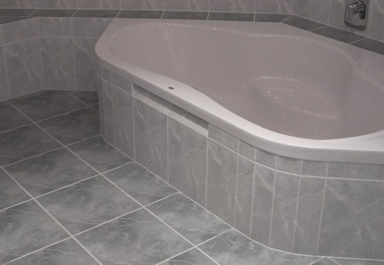Wykończona łazienka z narożną wanną obudowaną szarymi płytkami imitującymi marmur, podłoga wyłożona identycznymi płytkami, chromowana bateria widoczna w rogu.