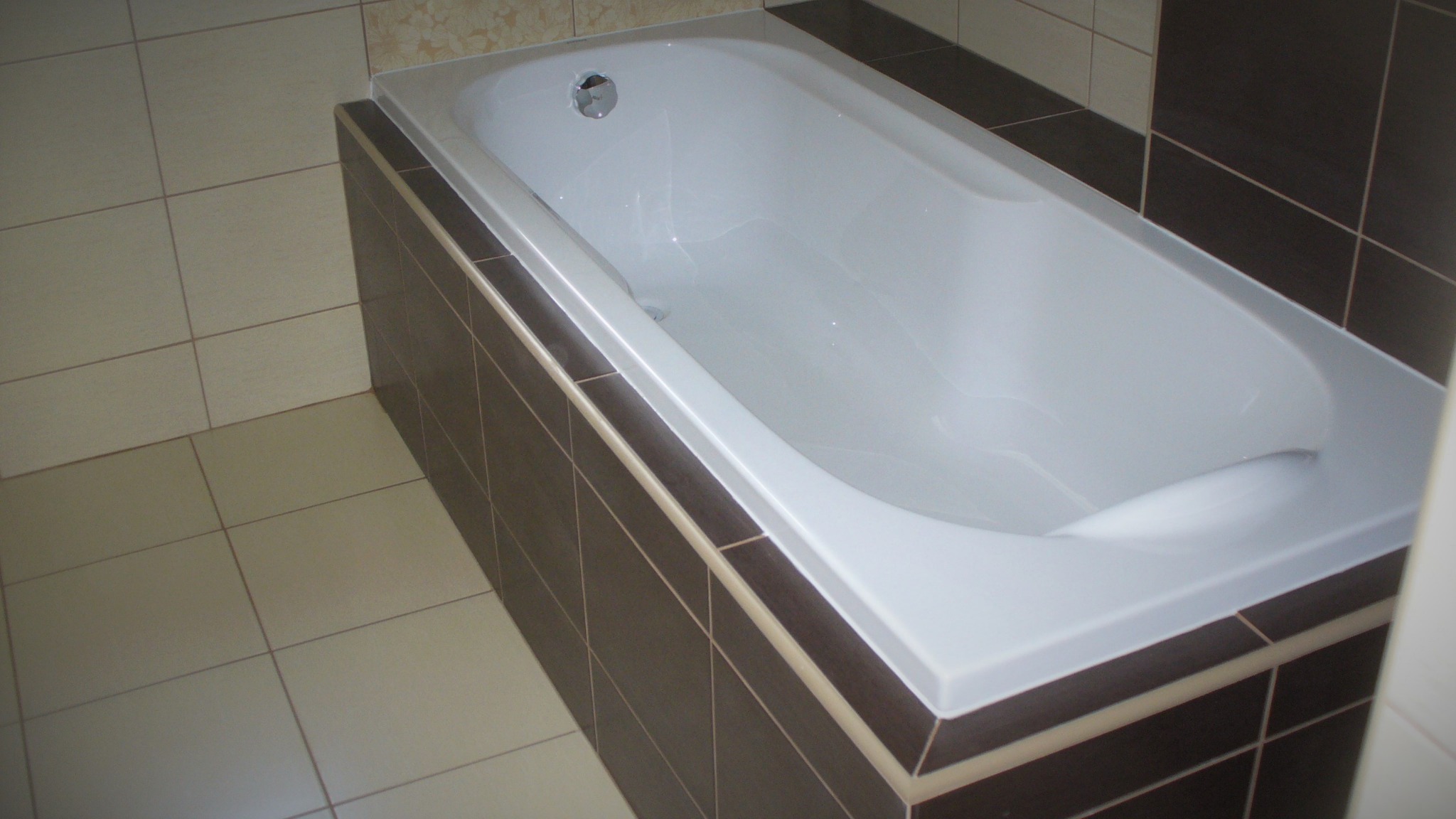 Wykończona łazienka z białą wanną obudowaną ciemnobrązowymi płytkami, jasne płytki podłogowe, widok z góry.