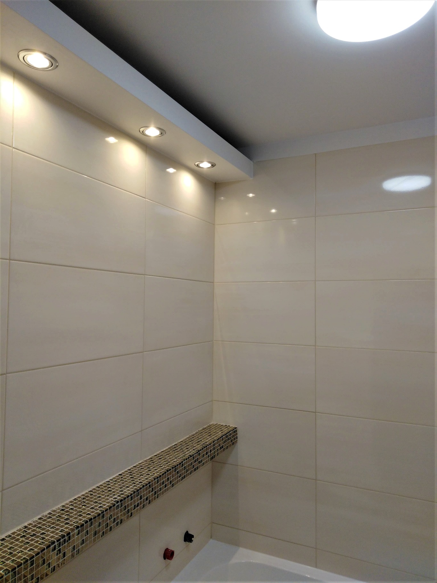 Wykończona łazienka z beżowymi, błyszczącymi płytkami, mozaikowym pasem dekoracyjnym i oświetleniem punktowym w suficie podwieszanym.