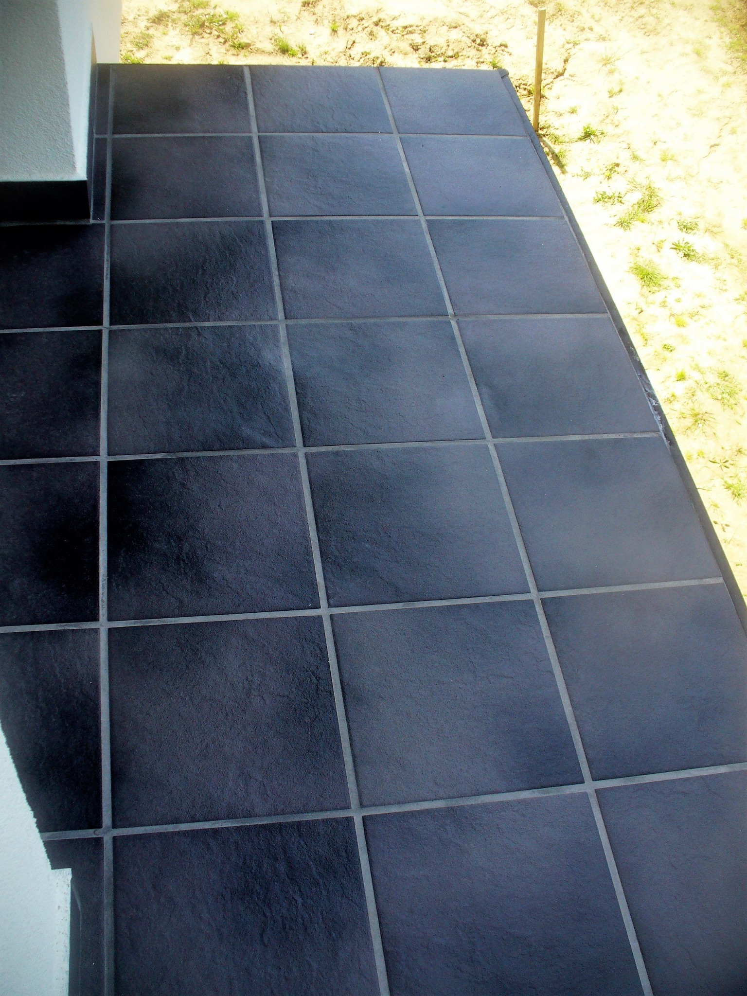 Ciemne, kwadratowe płytki gresowe ułożone na zewnątrz, z widocznymi fugami i fragmentem ściany w tle.
