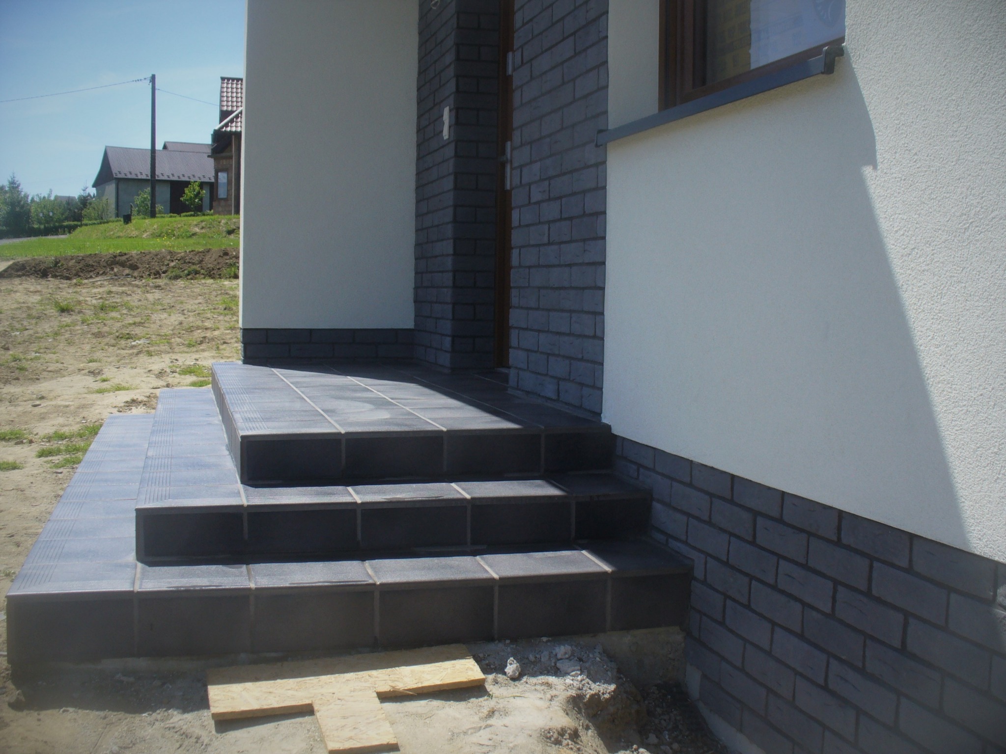 Wykończone płytkami schody wejściowe do domu z elewacją w odcieniach szarości, widoczne deski na ziemi.