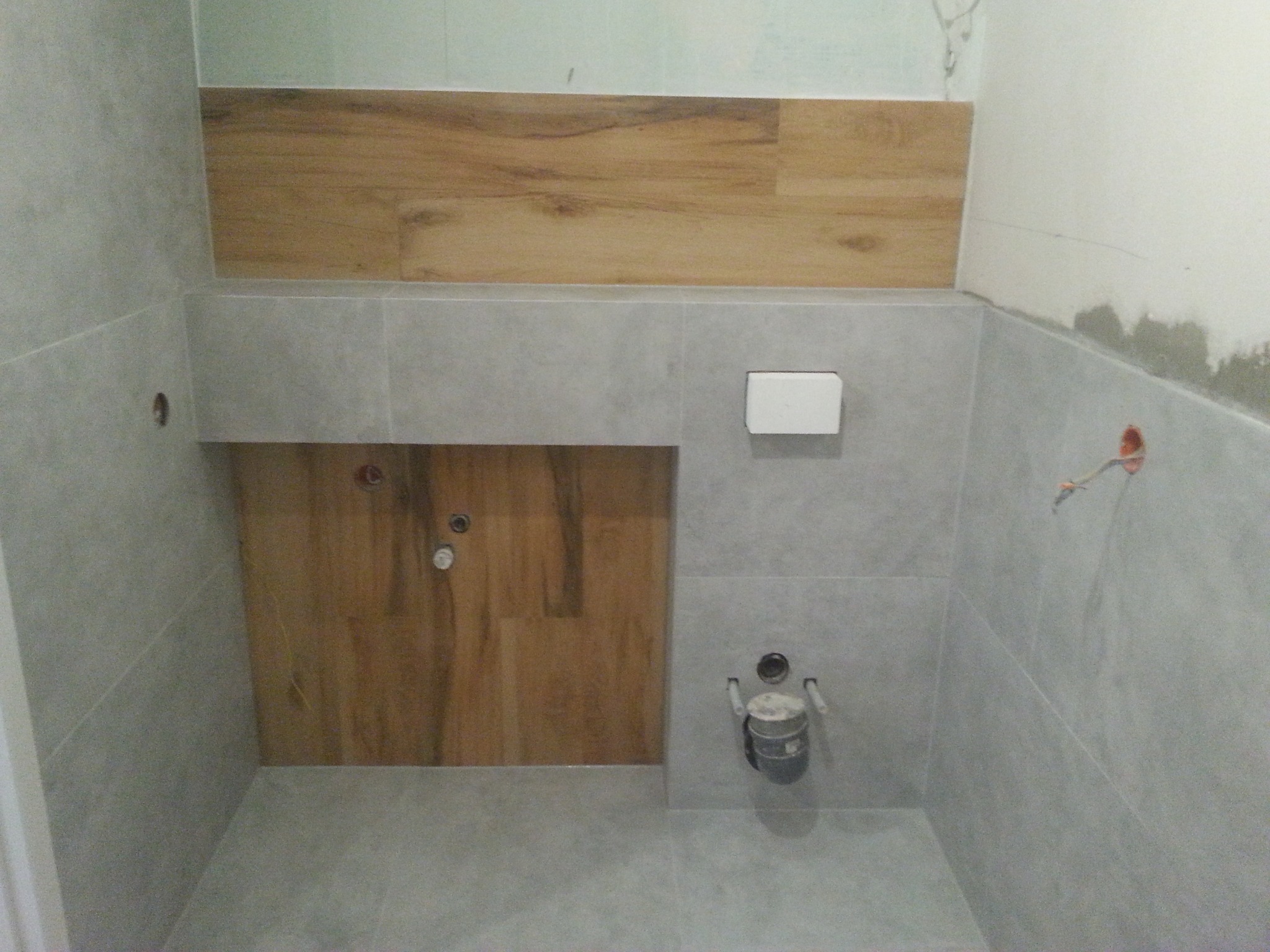 Półka z imitacji drewna nad szarą ścianą z płytek w łazience w trakcie remontu, widoczne instalacje hydrauliczne.