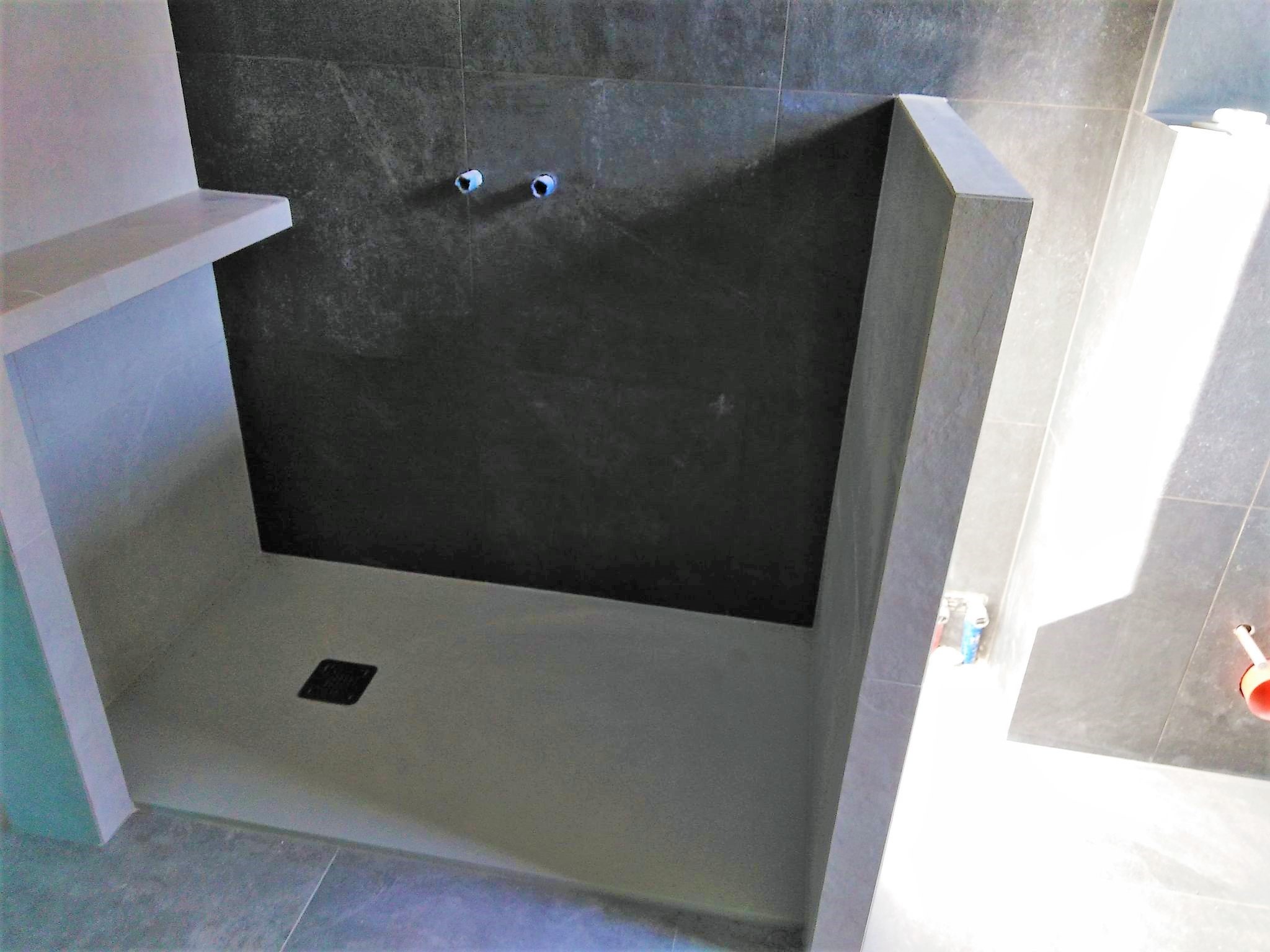 Wykończony brodzik z ciemnymi płytkami na ścianie, widoczne przyłącza wodne, odpływ liniowy i półka wnękowa