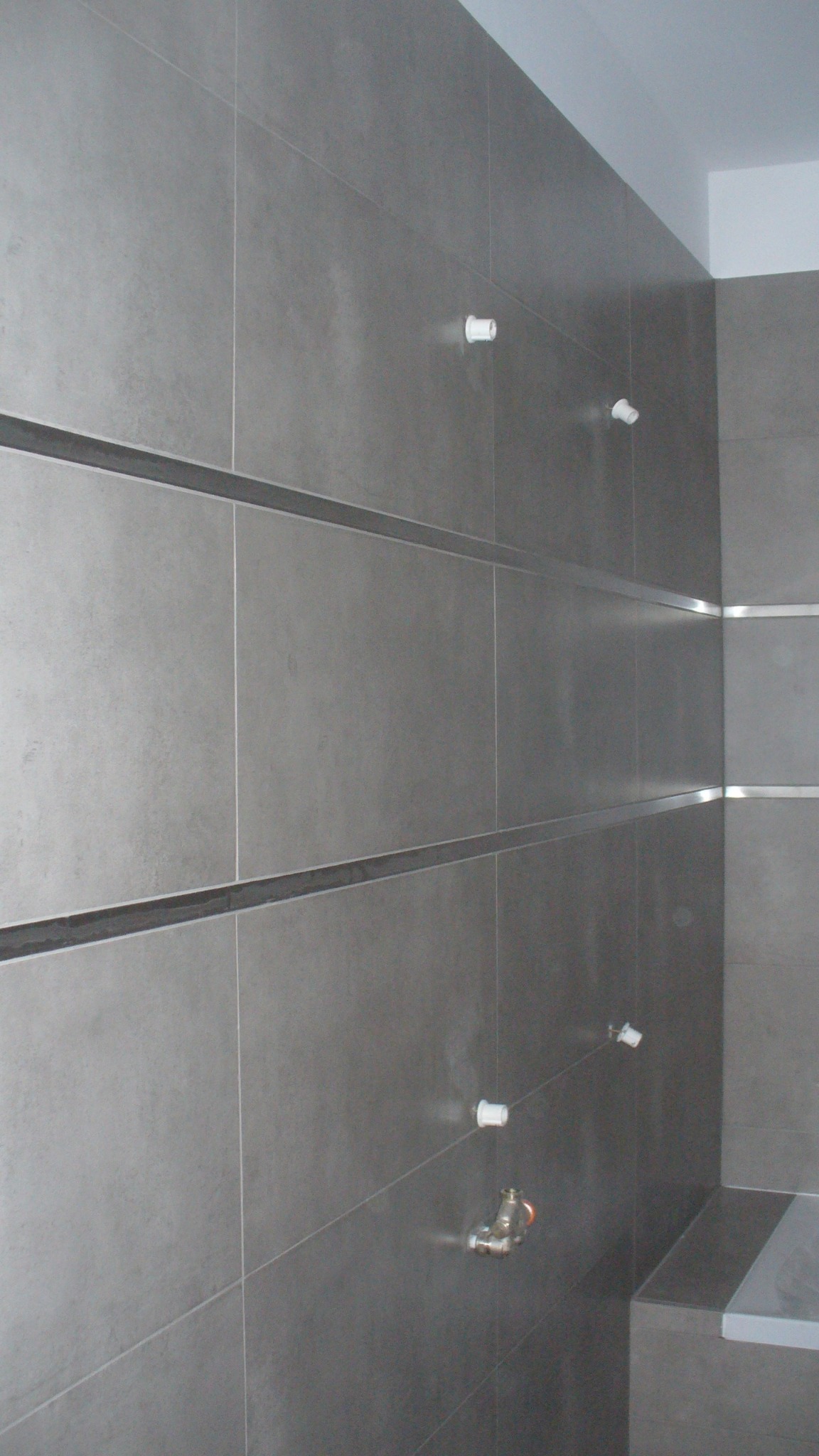 Szare, matowe płytki ceramiczne w łazience, z widocznymi otworami na baterie i dyskretnymi, poziomymi listwami dekoracyjnymi.