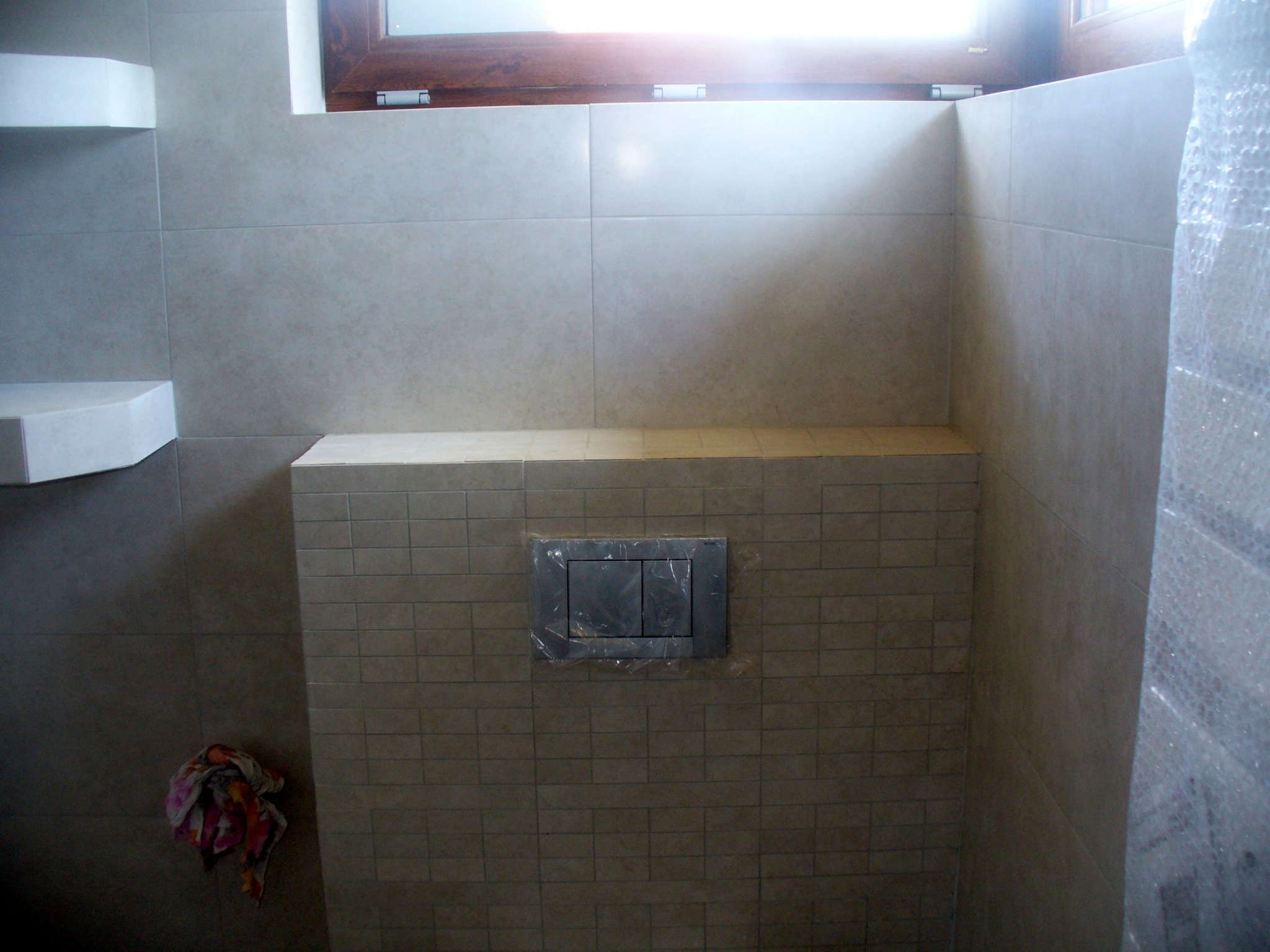 Wykończona łazienka z beżowymi płytkami, widoczny fragment okna, półka narożna i przycisk spłuczki zabezpieczony folią.