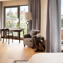 Elegancka sypialnia z widokiem na Wawel, szare zasłony z geometrycznym wzorem, drewniane biurko i fotel, lampa podłogowa i stolik nocny z dekoracyjnym wazonem.