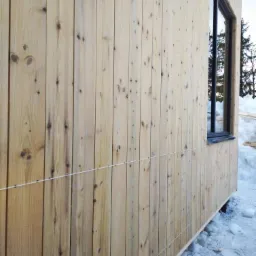 Ściana budynku obita pionowymi, jasnymi deskami z widocznymi sękami, z oknem w czarnej ramie i poziomą linką wyznaczającą poziom, otoczona śniegiem.