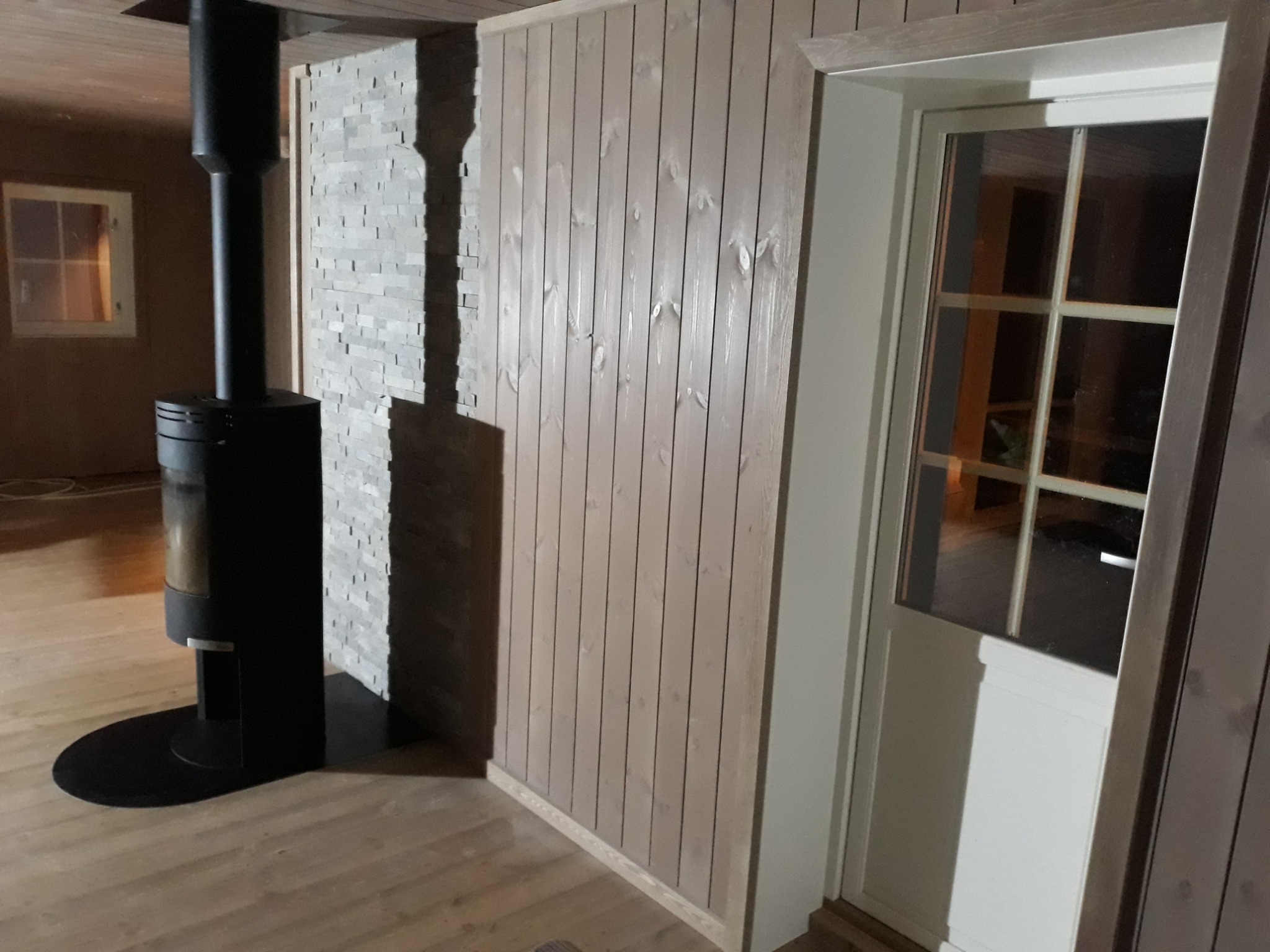Wnętrze pokoju z drewnianą boazerią pionową pomalowaną na jasny kolor, fragmentem ściany wyłożonej dekoracyjnym kamieniem, czarnym piecem wolnostojącym i białymi drzwiami z szybami.