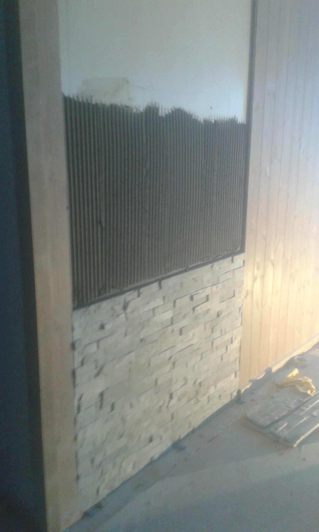 Ściana z częściowo ułożoną okładziną z kamienia dekoracyjnego, widoczne ślady kleju na górnej części ściany i fragment drewnianej boazerii obok.