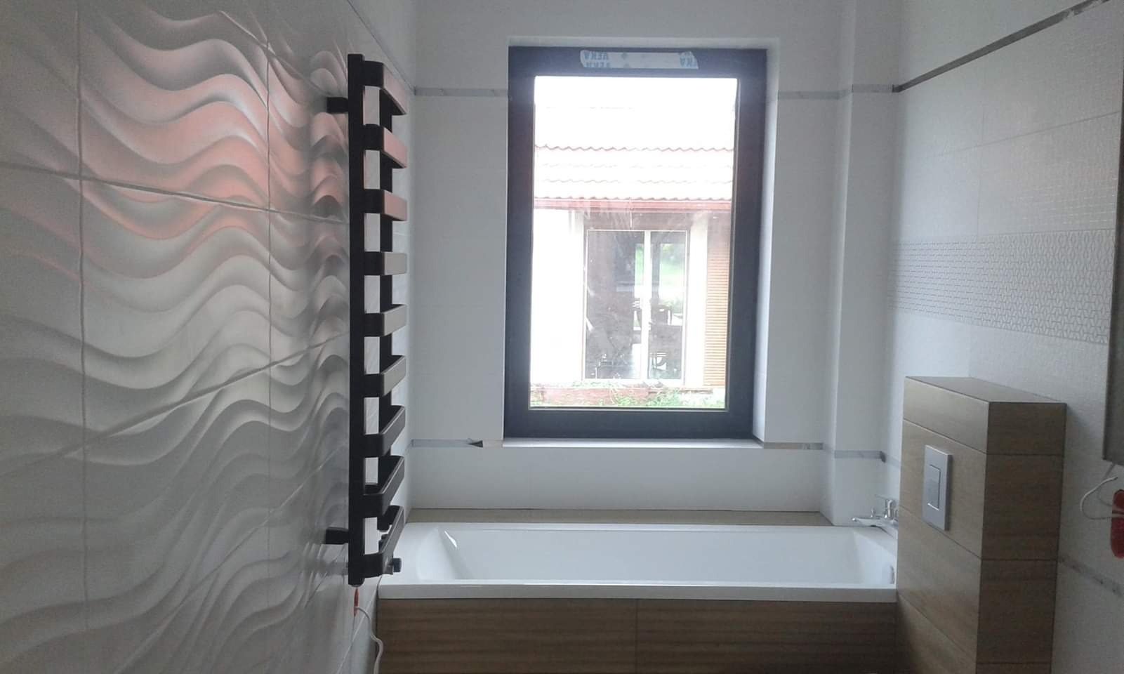 Nowoczesna łazienka z białą wanną pod oknem, grzejnikiem drabinkowym na ścianie z trójwymiarowymi płytkami w kształcie fal i zabudową WC z drewnopodobnym wykończeniem.