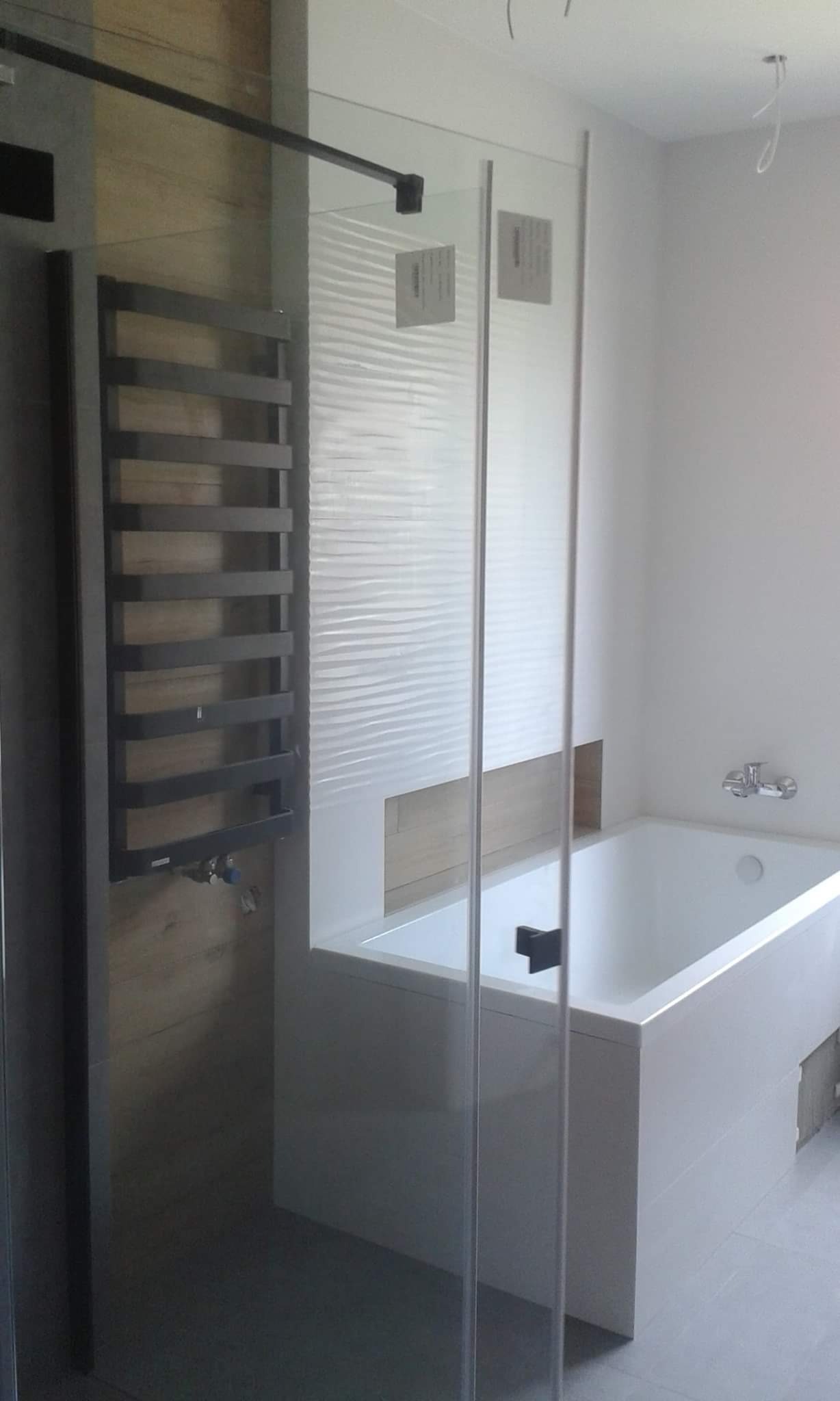 Nowoczesna łazienka z wanną obudowaną białą płytą, szklanym parawanem nawannowym i grzejnikiem drabinkowym w kolorze grafitowym na ścianie wyłożonej drewnopodobnymi płytkami.