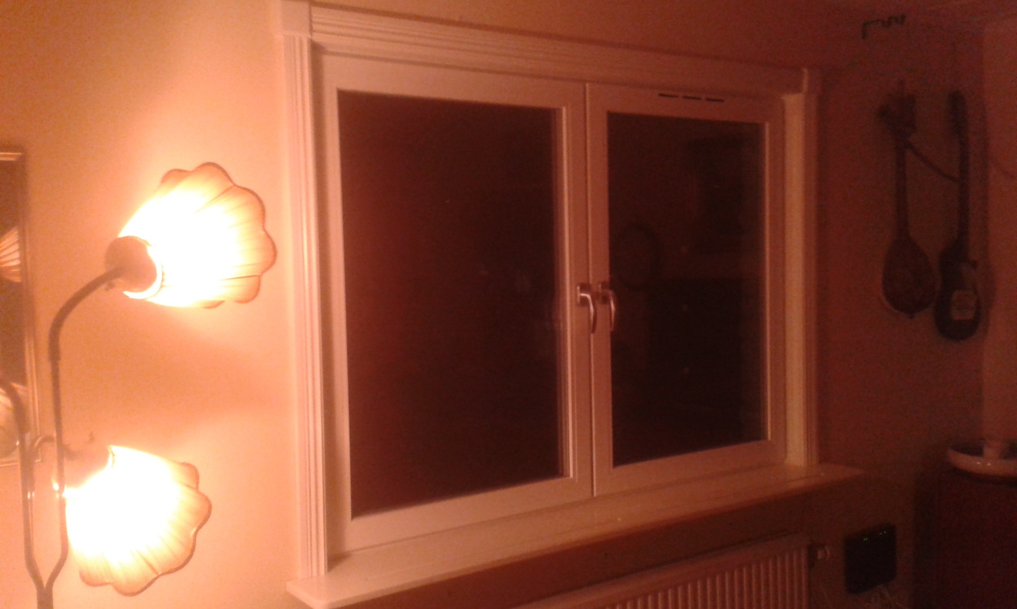 Białe, dwuskrzydłowe okno PCV z dekoracyjną ramą, oświetlone ciepłym światłem lampy stojącej w kształcie kwiatu, z widocznymi instrumentami muzycznymi wiszącymi na ścianie w tle.