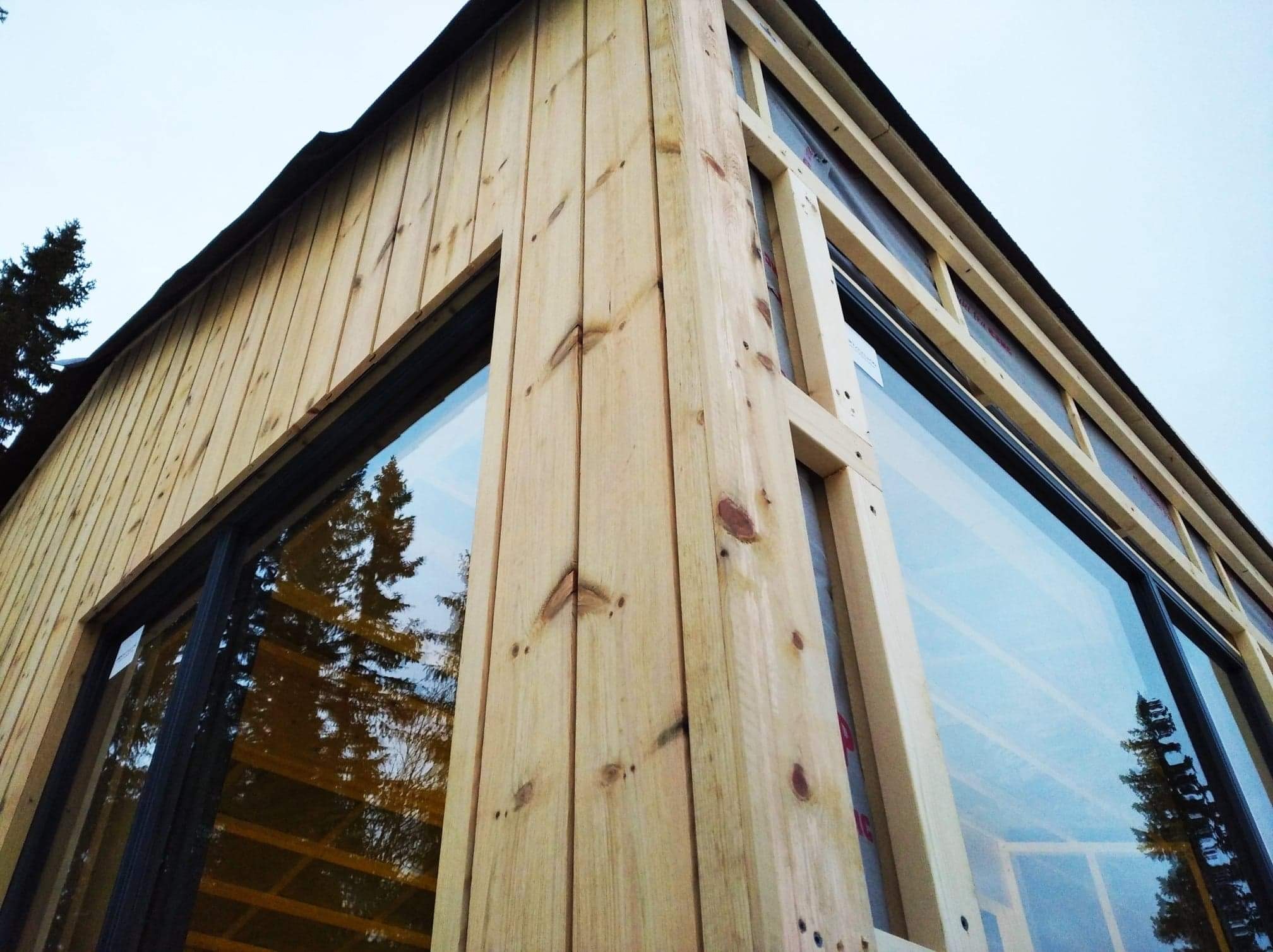 Narożnik budynku w trakcie budowy, widoczne drewniane deski elewacyjne i okna z czarnymi ramami, w których odbijają się drzewa.
