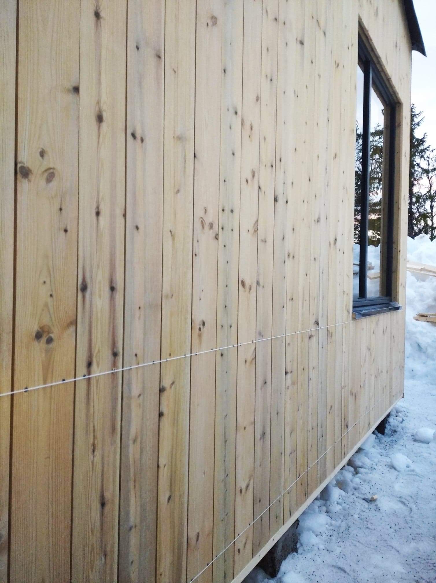 Ściana budynku obita pionowymi, jasnymi deskami z widocznymi sękami, z oknem w czarnej ramie i poziomą linką wyznaczającą poziom, otoczona śniegiem.
