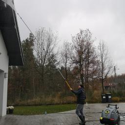 Pro-Jet - Mężczyzna w czapce i kamizelce czyści rynnę wysokociśnieniową lancą teleskopową, obok stoi myjka ciśnieniowa, w tle drzewa i dom.