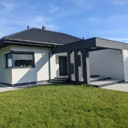 Nowoczesny parterowy dom z płaskim dachem, białymi ścianami i antracytowymi elementami, takimi jak okna, obramowania dachu i wiata garażowa, na tle błękitnego nieba.