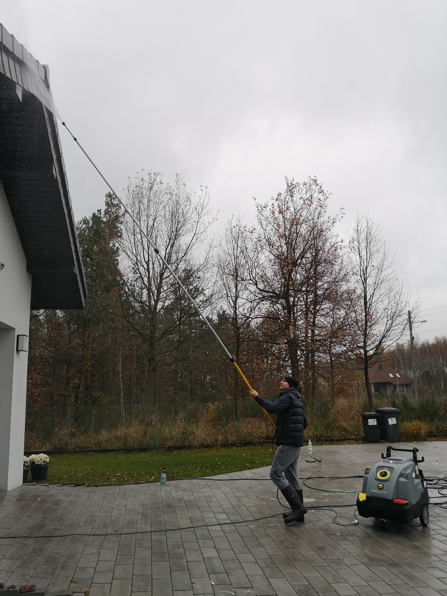 Mężczyzna w czapce i kamizelce czyści rynnę wysokociśnieniową lancą teleskopową, obok stoi myjka ciśnieniowa, w tle drzewa i dom.
