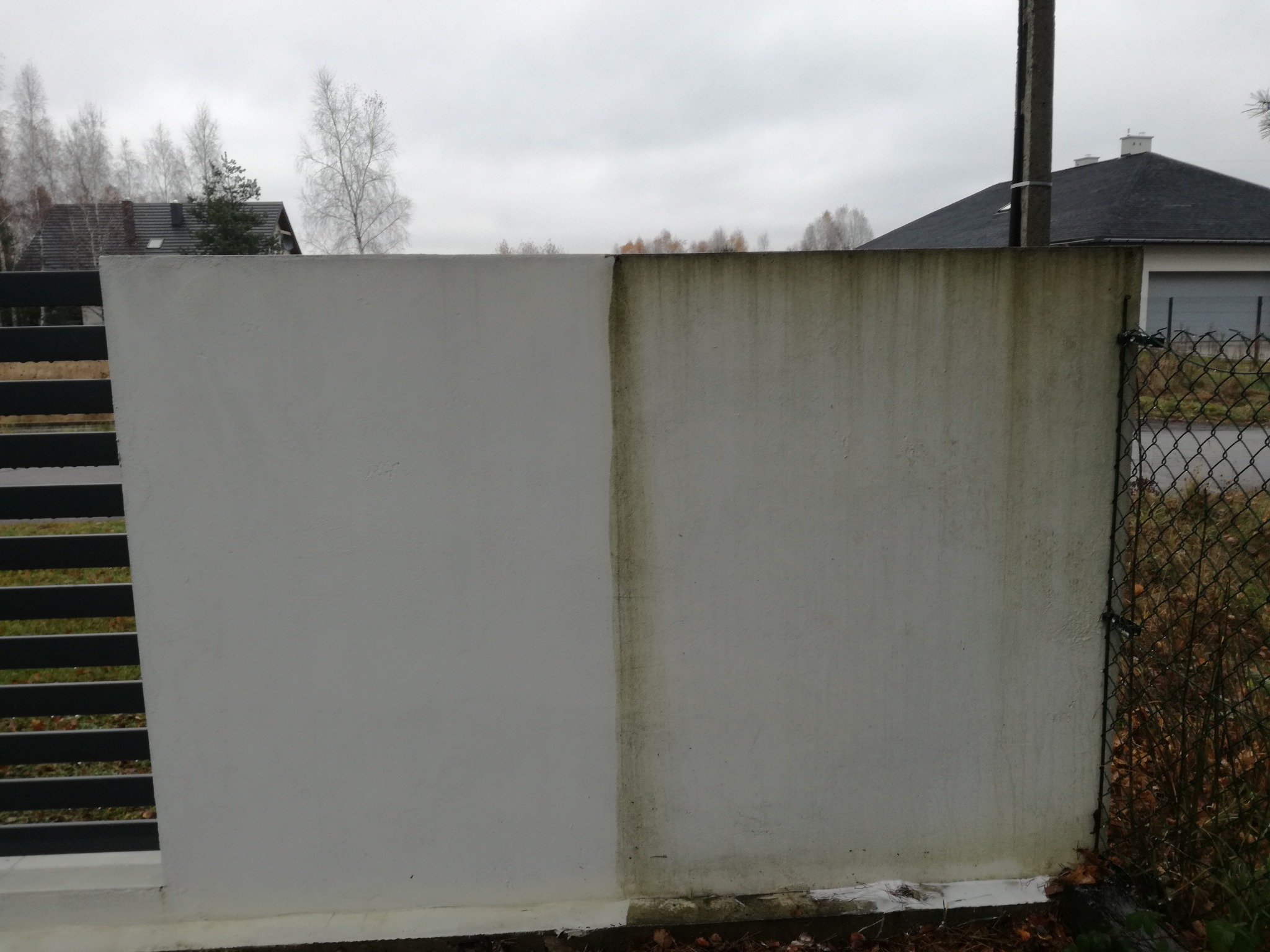 Fragment ściany zewnętrznej, na której widoczny jest efekt czyszczenia, po lewej stronie ściana jest czysta i pomalowana na biało, a po prawej brudna i pokryta nalotem. W tle widoczny dom i drzewa.