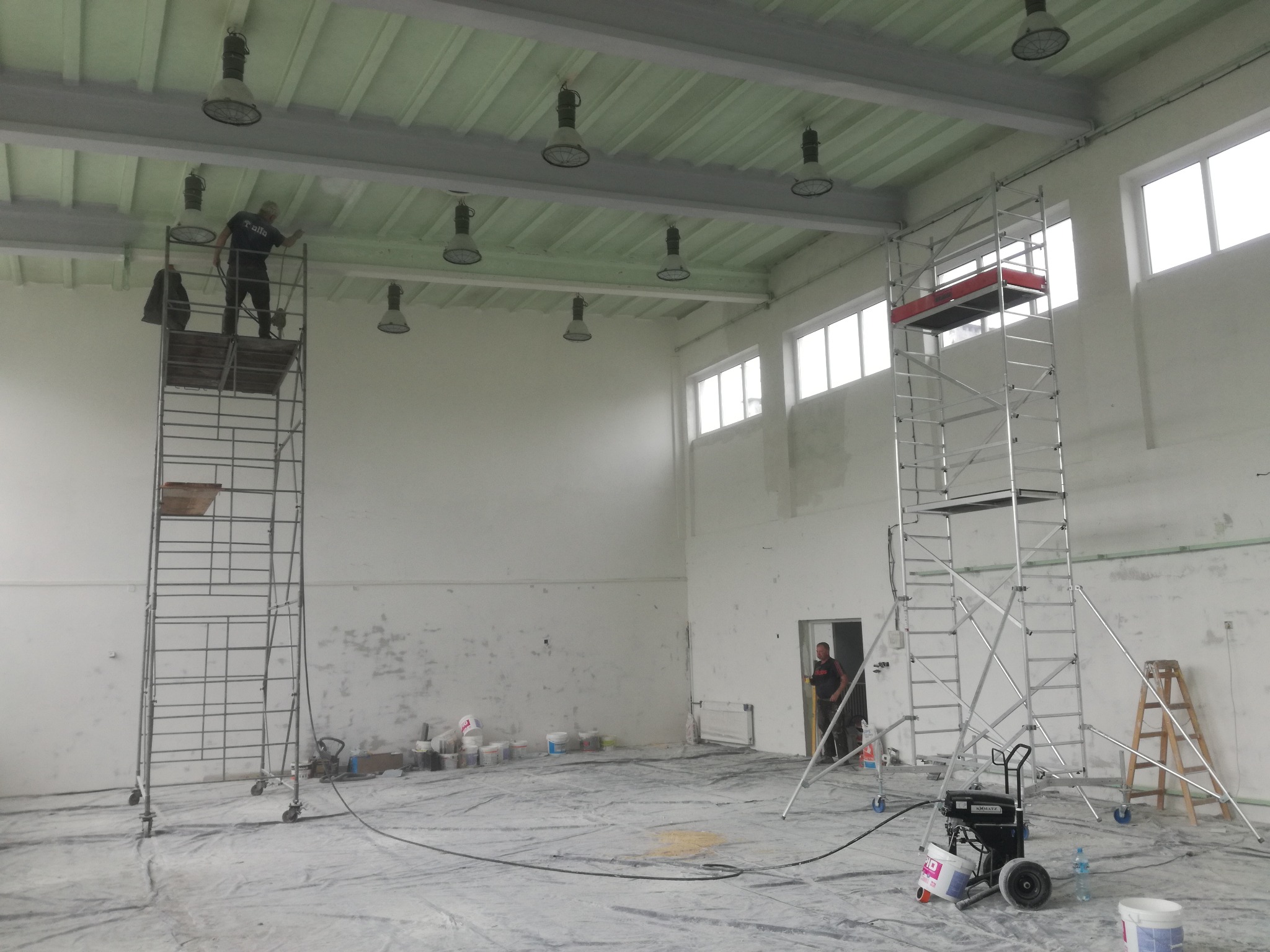 Malowanie sufitu w hali przemysłowej; pracownik na rusztowaniu, widoczne puszki z farbą, agregat malarski i zabezpieczona folią podłoga.