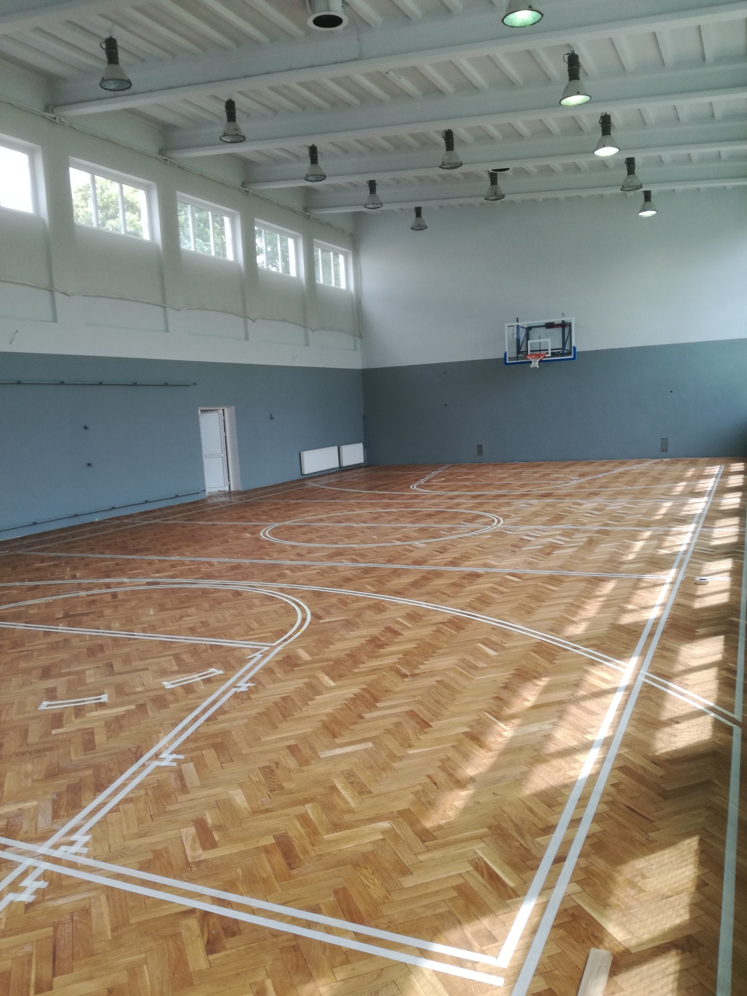 Wnętrze sali gimnastycznej z parkietem ułożonym w jodełkę, świeżo pomalowanymi liniami boiska i koszem do koszykówki na ścianie.