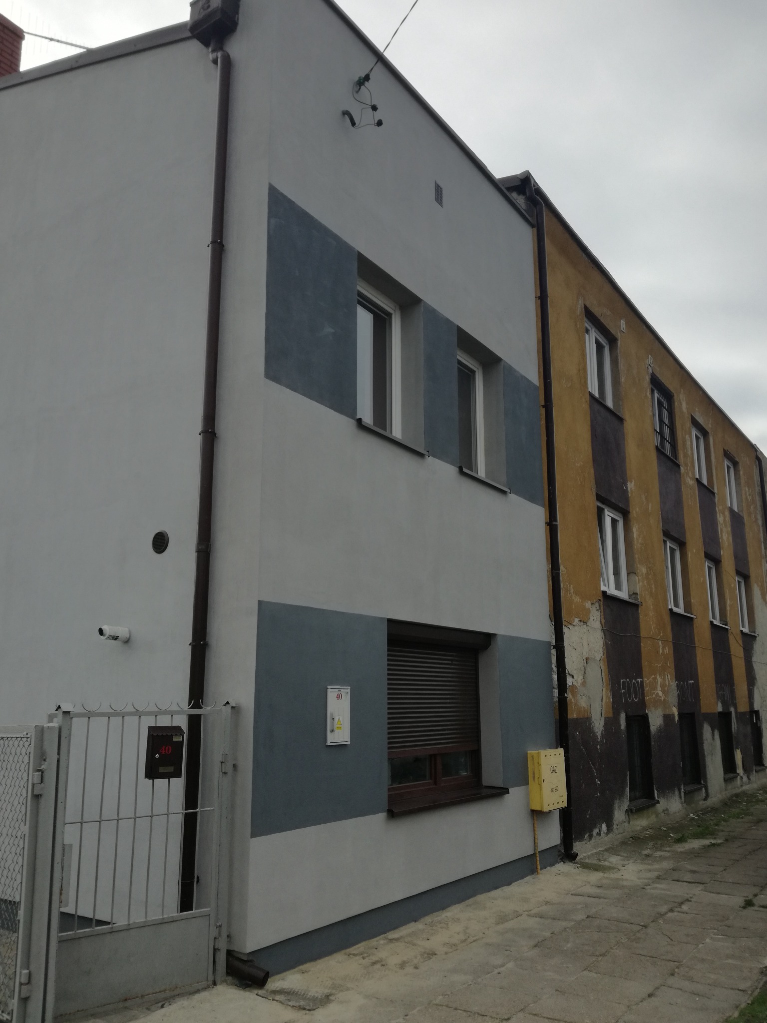 Dwukondygnacyjny budynek z częściowo odnowioną elewacją w odcieniach szarości i błękitu, kontrastujący z fragmentem budynku ze starą, zniszczoną, żółto-brązową elewacją z widocznymi ubytkami...