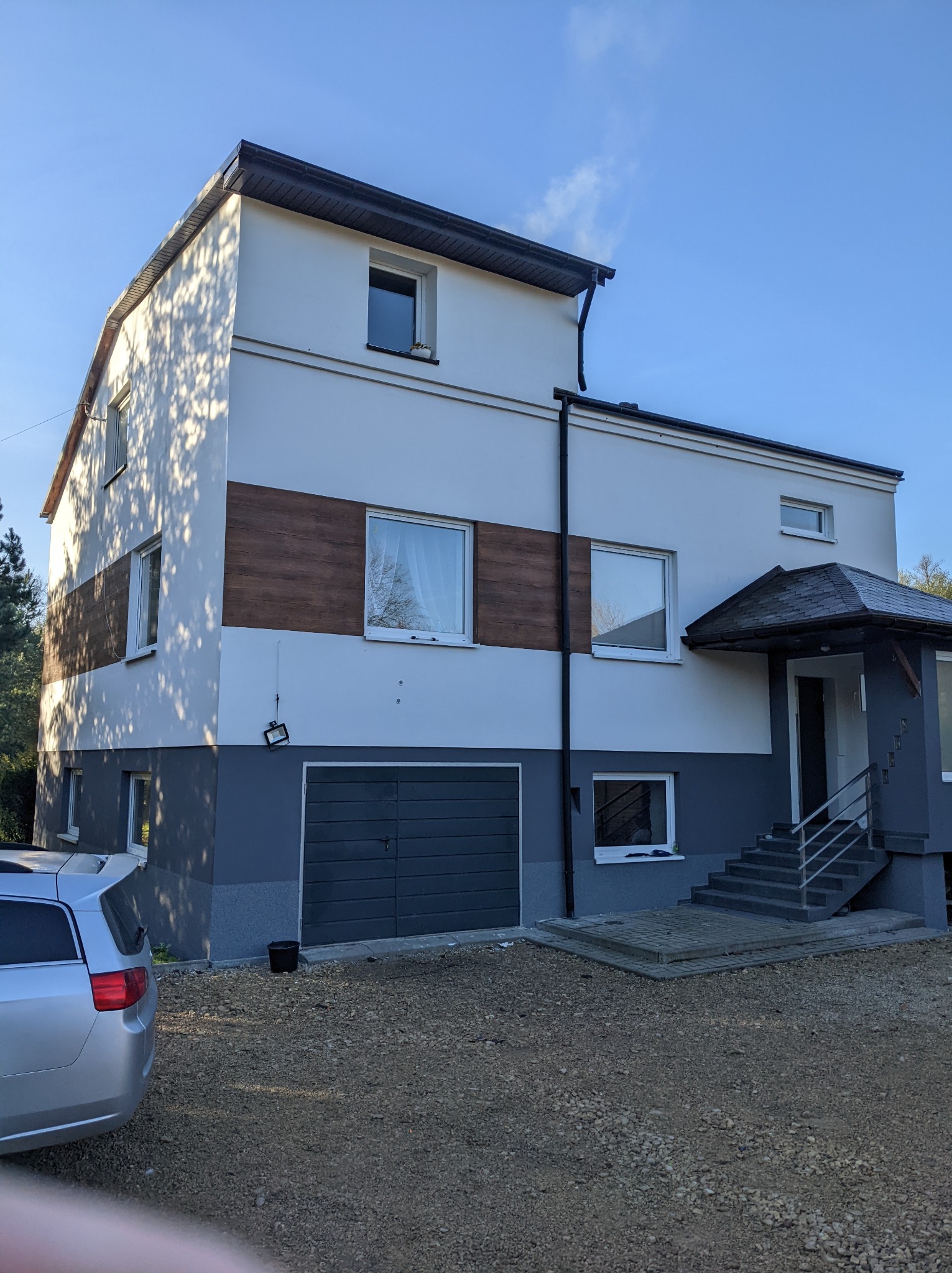 Dwukondygnacyjny dom z szarą elewacją w dolnej części i białym tynkiem powyżej, z poziomym pasem imitującym drewno, garażem i schodami wejściowymi pod zadaszeniem.