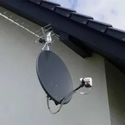 Antena satelitarna i antena naziemna zamontowane na ścianie domu z ciemną dachówką i oknami z brązowymi okiennicami, na tle błękitnego nieba z chmurami.