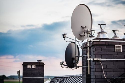 Dwie anteny satelitarne zamontowane na kominie z cegły, widok z dołu, na tle niebieskiego nieba z chmurami.