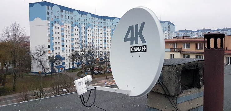 Biała antena satelitarna 4K Canal+ zamontowana na dachu budynku z widokiem na osiedle bloków mieszkalnych w tle.