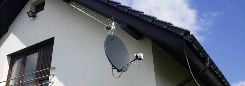 Antena satelitarna i antena naziemna zamontowane na ścianie domu z ciemną dachówką i oknami z brązowymi okiennicami, na tle błękitnego nieba z chmurami.