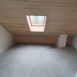 Poddasze z drewnianą boazerią na skosach i suficie, okno dachowe oraz surowa betonowa podłoga w trakcie prac wykończeniowych.