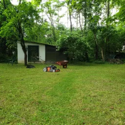 Pomarańczowa kosiarka spalinowa na trawniku przed białym budynkiem gospodarczym, w tle taczka i drzewa.