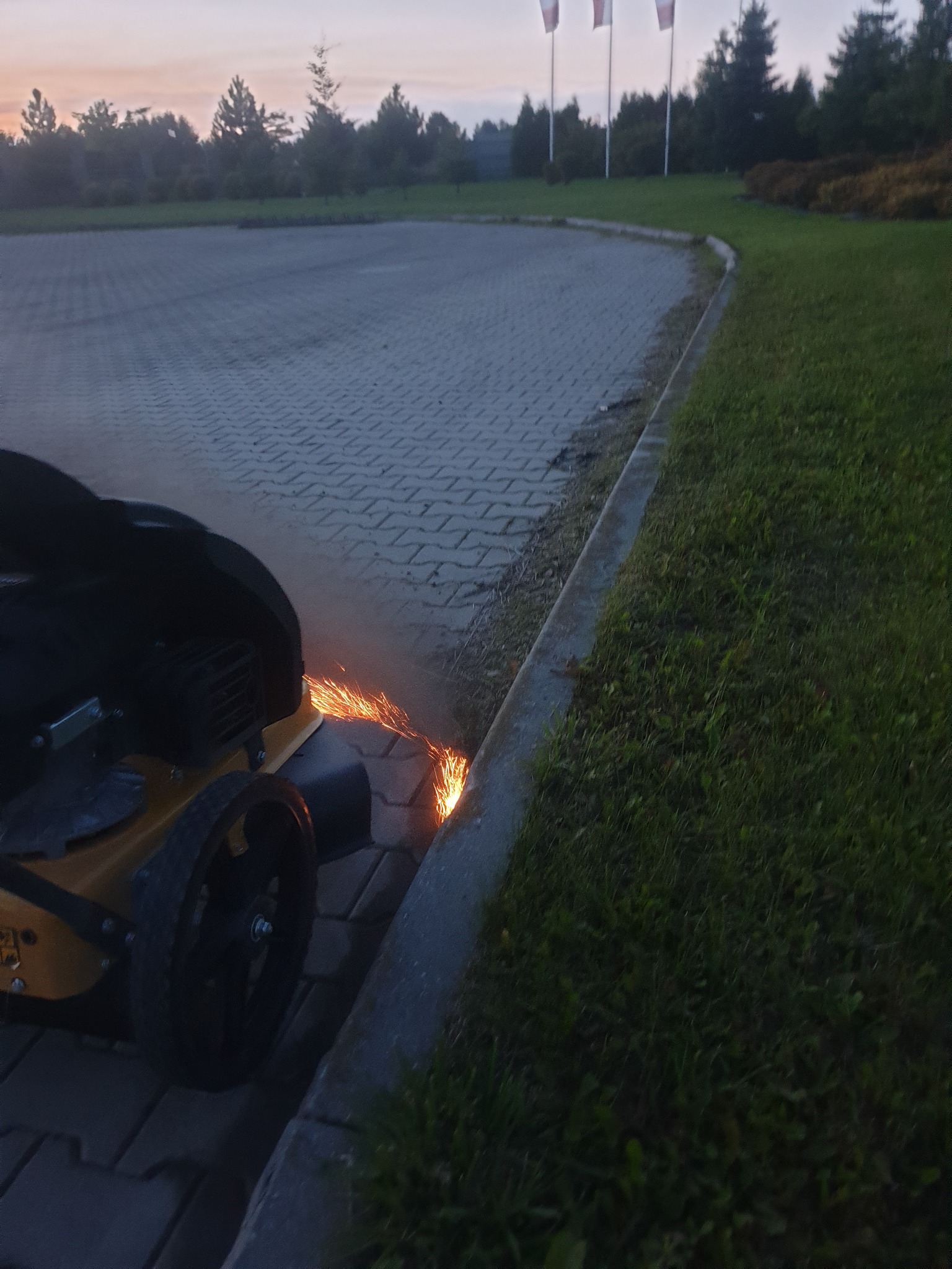 Żółta kosiarka spalinowa z osłoną tnącą, iskrząca podczas przycinania krawężnika trawnika na parkingu z kostki brukowej o zmierzchu.