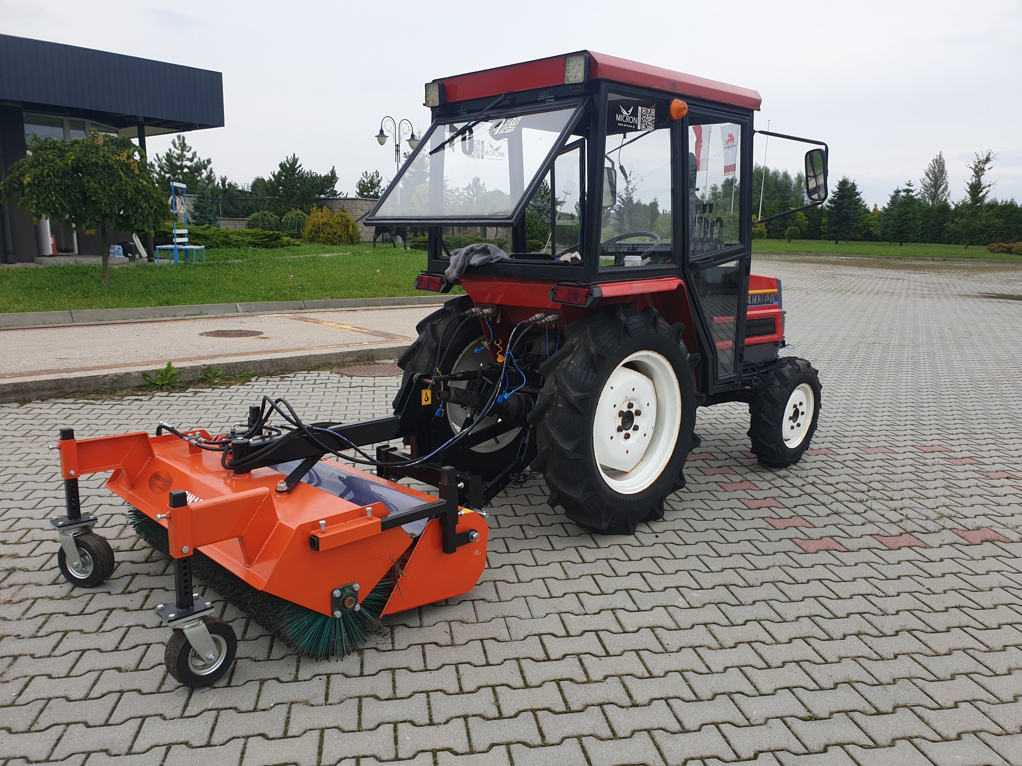 Czerwony traktor z kabiną i pomarańczową szczotką do zamiatania na kostce brukowej, widok z tyłu.