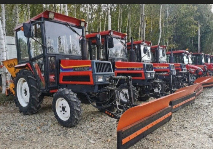 Rząd czerwonych traktorów marki Yanmar z zamontowanymi pługami do odśnieżania, zaparkowanych na żwirowym placu, z widocznymi drzewami w tle.
