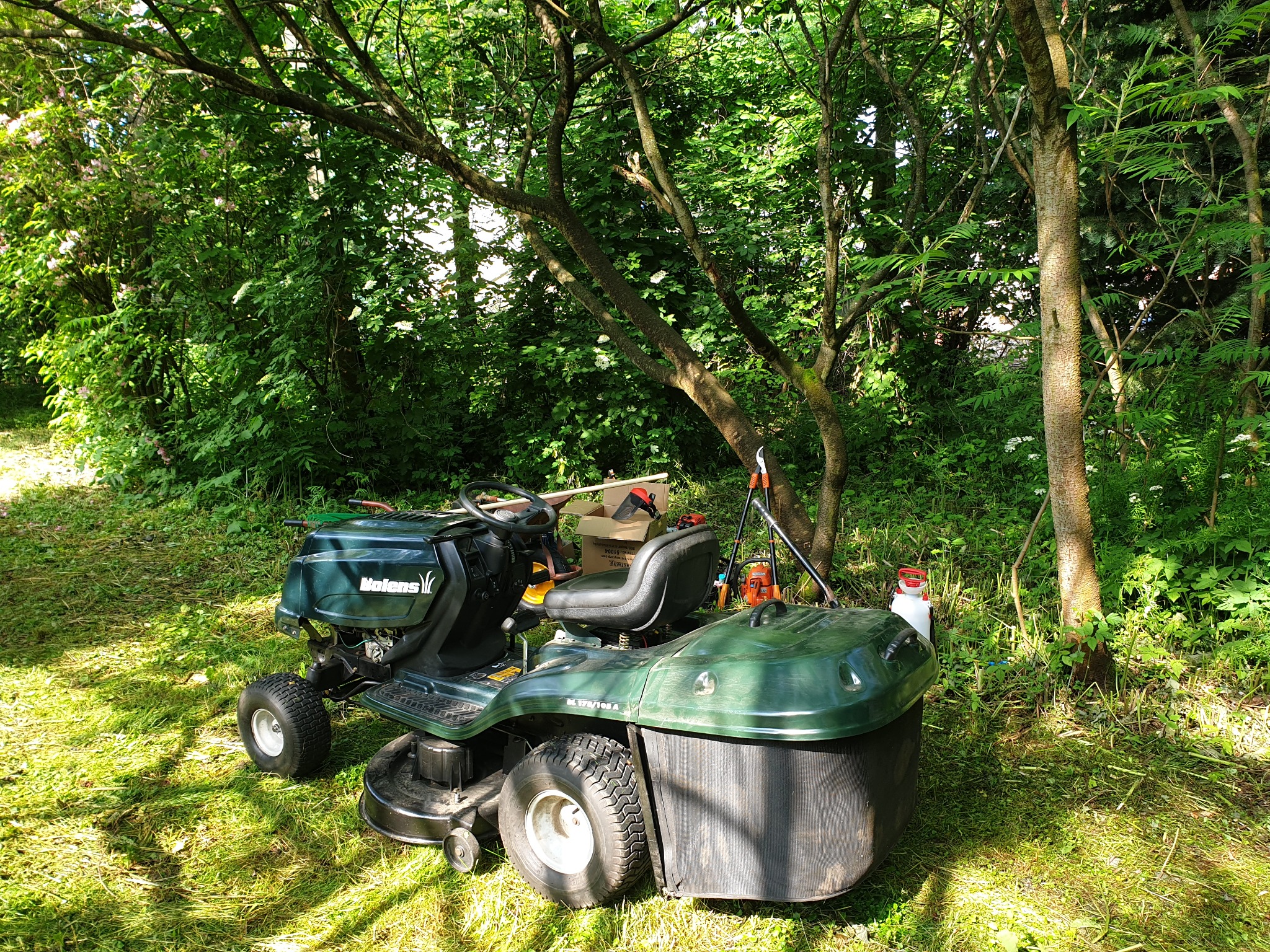 Zielony traktorek do koszenia trawy marki MTD Bolens zaparkowany na trawniku w cieniu drzew, obok widoczne narzędzia ogrodnicze i pojemnik na paliwo.