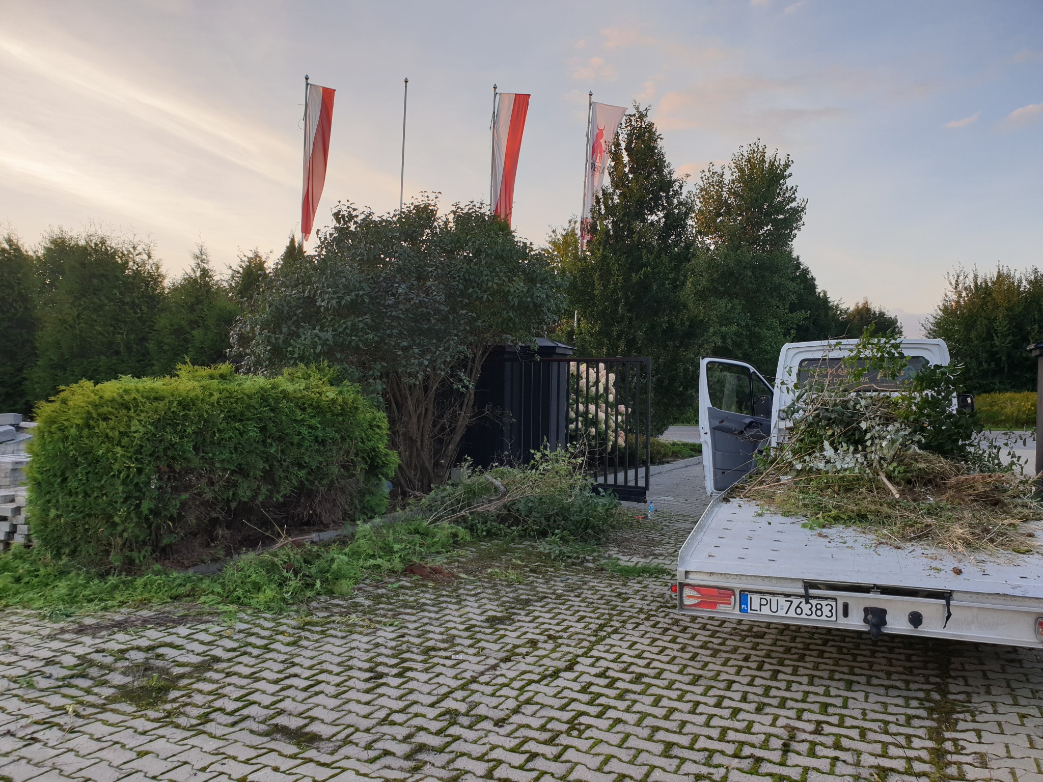 Przyczepa z otwartymi drzwiami wypełniona gałęziami i liśćmi po wycince drzew, obok przycięty krzew, na tle trzy flagi na masztach.
