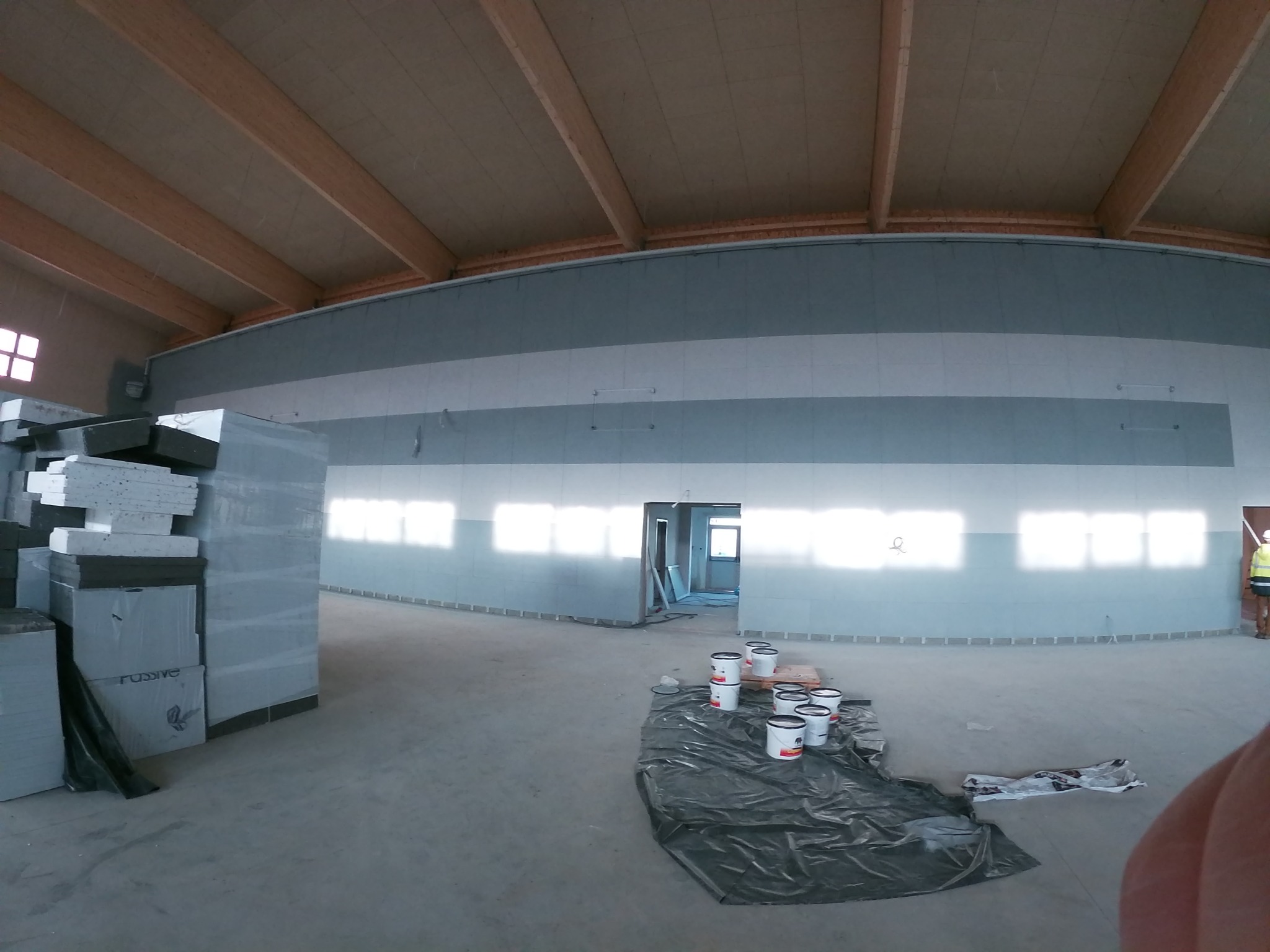 Wnętrze hali w trakcie remontu, widoczne puszki z farbą na folii, płyty kartonowo-gipsowe w opakowaniach oraz ściany przygotowane do malowania z drewnianą konstrukcją dachu.