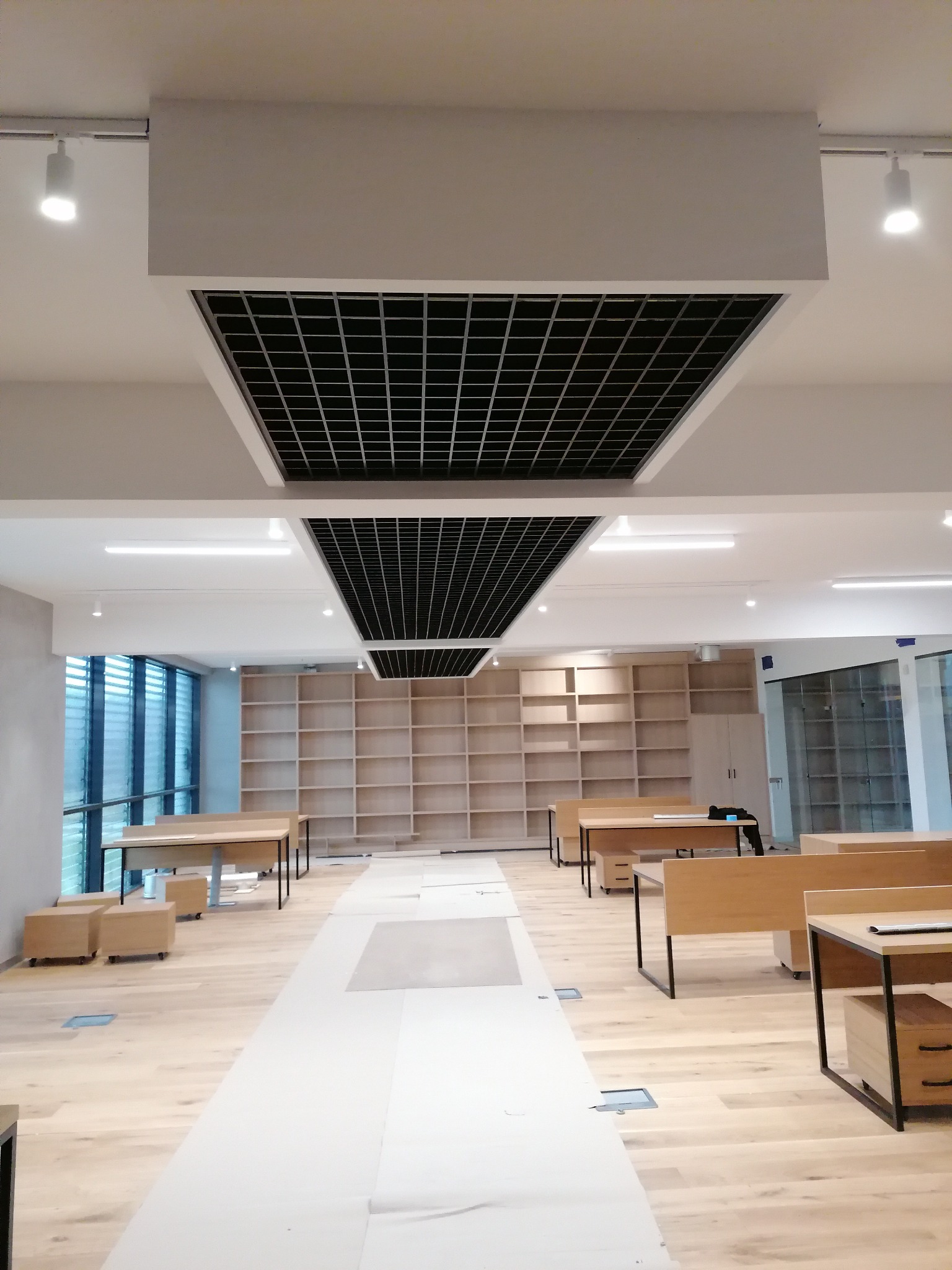 Wnętrze biurowe w trakcie wykańczania, z widocznymi podwieszanymi sufitami rastrowymi, drewnianą podłogą zabezpieczoną folią, biurkami i regałami w tle.