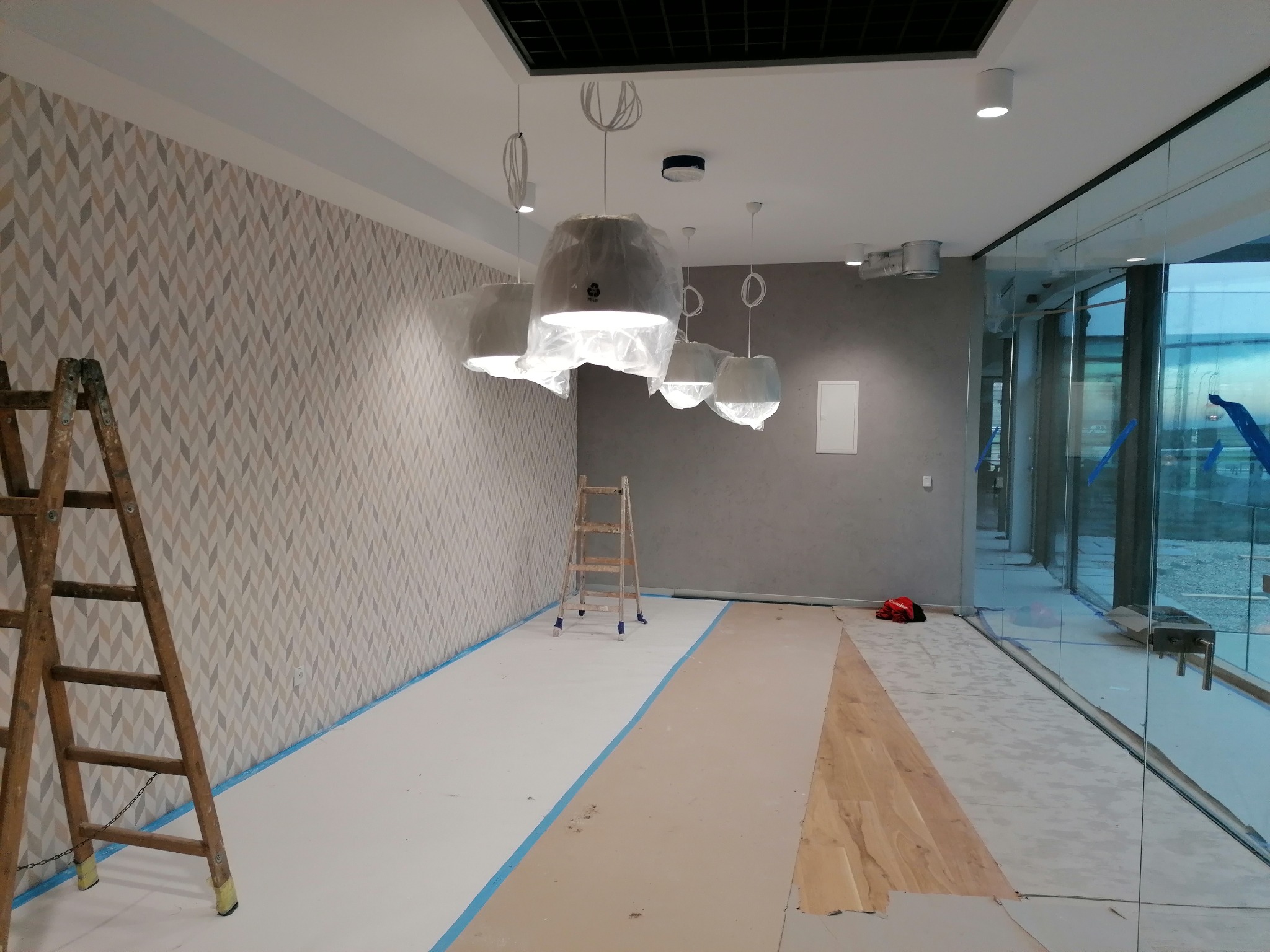 Wnętrze w trakcie prac wykończeniowych: zabezpieczona podłoga, drabina oparta o ścianę z wzorzystą tapetą, lampy wiszące osłonięte folią, ściana pomalowana na szaro, duże okno z widokiem na zewnątrz.