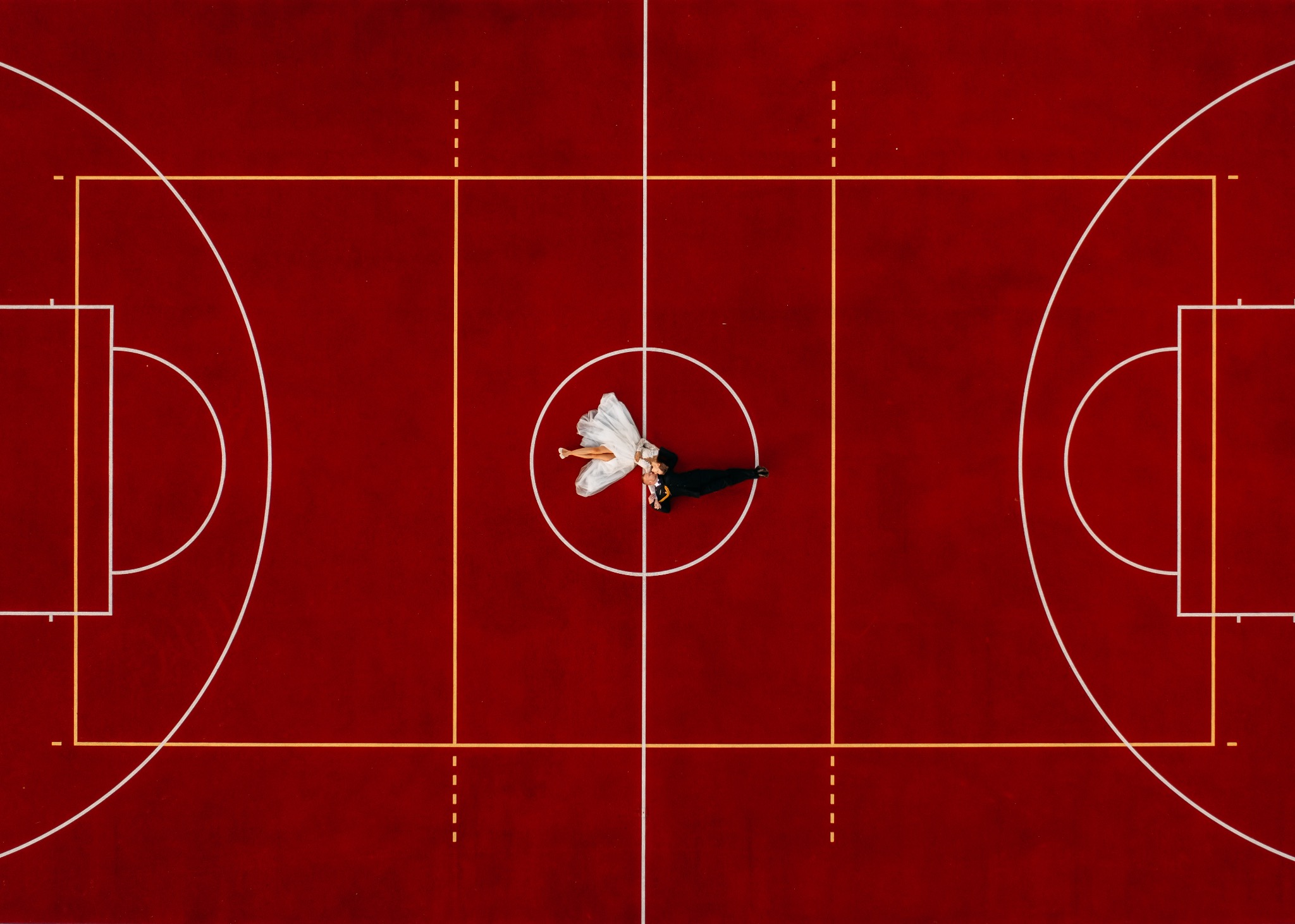 Para młoda leżąca na środku boiska do koszykówki, widok z góry, panna młoda w białej sukni, pan młody w czarnym garniturze, czerwone tło boiska z białymi i żółtymi liniami.