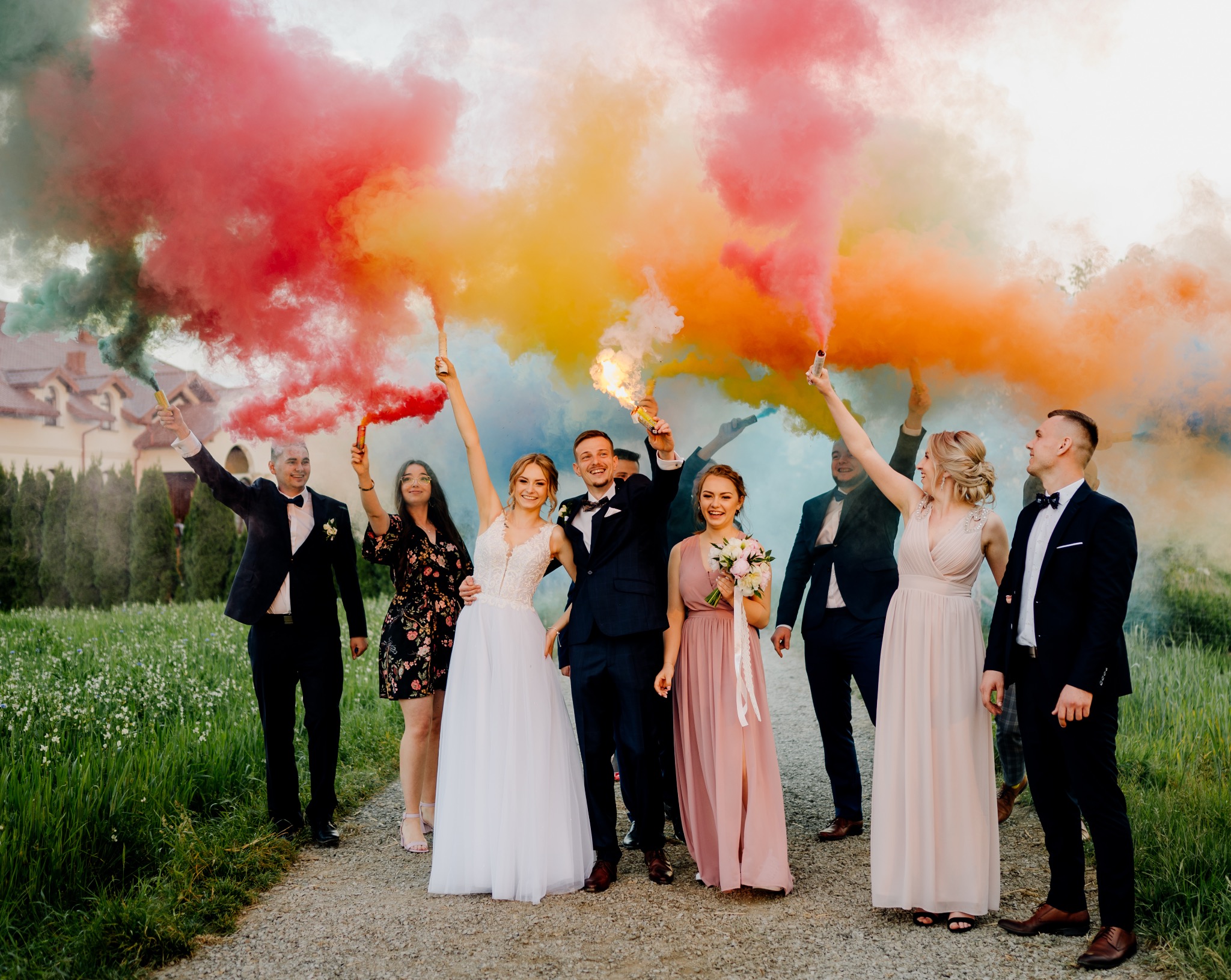 Para młoda z gośćmi weselnymi świętuje z kolorowymi racami dymnymi na tle zielonej łąki i domu z czerwonym dachem.