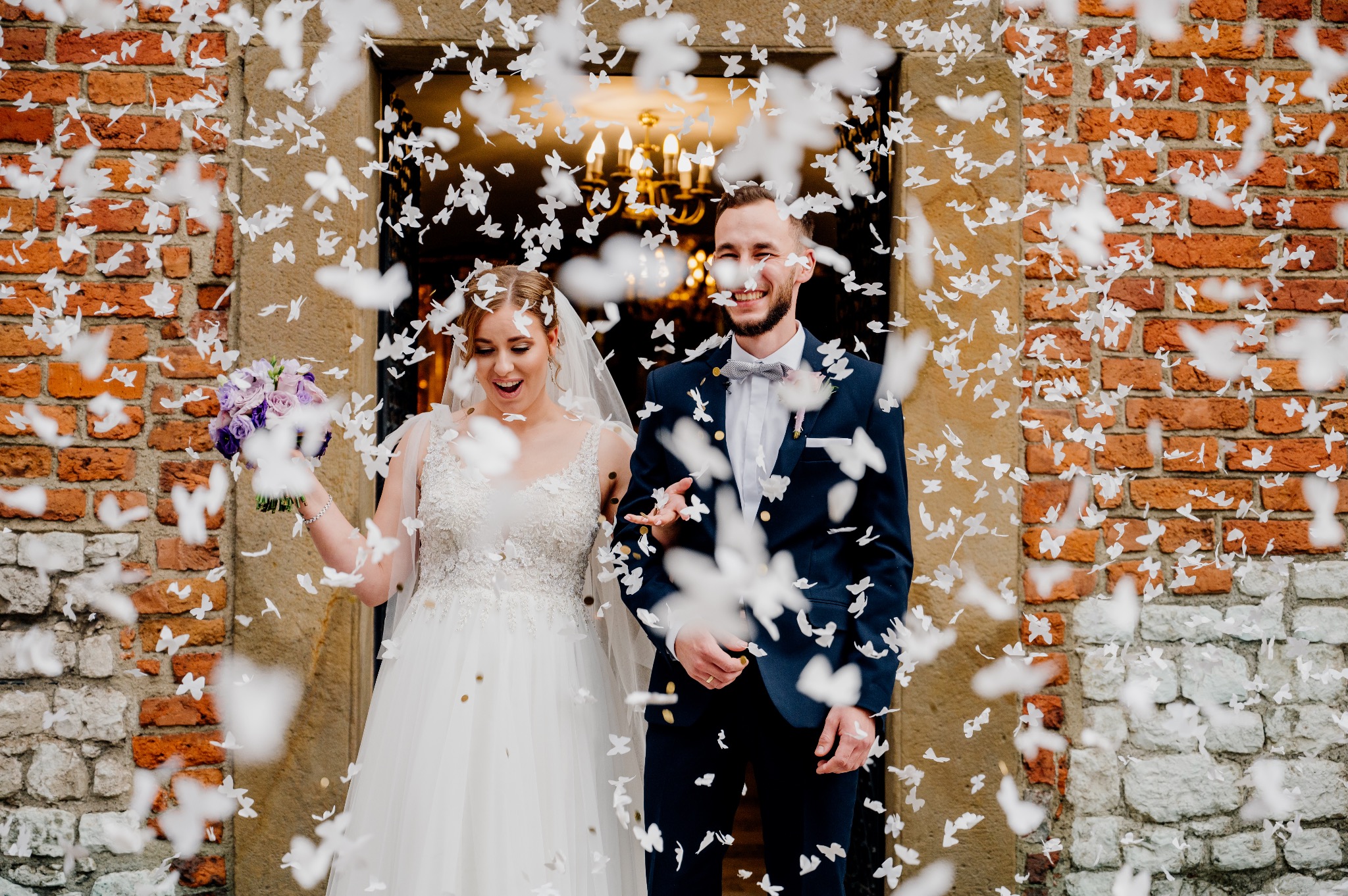 Uśmiechnięta para młoda wychodzi z budynku w deszczu białych konfetti w kształcie motyli, panna młoda trzyma bukiet fioletowych kwiatów.