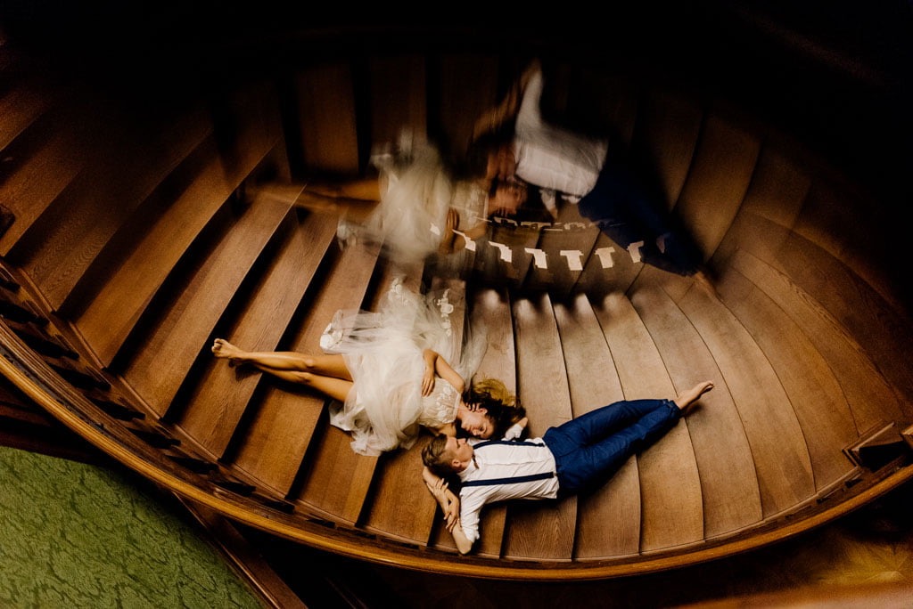 Para młoda leżąca na drewnianych kręconych schodach, ujęcie z góry z efektem rybiego oka, panna młoda w koronkowej sukni, pan młody w niebieskich spodniach i białej koszuli z szelkami.