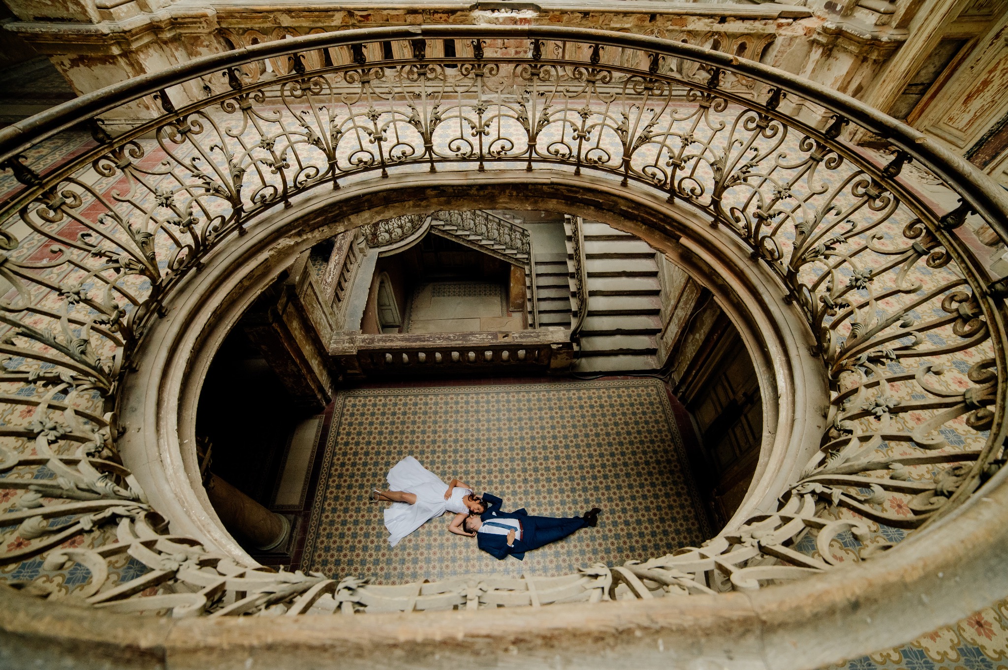 Para młoda leżąca na wzorzystej podłodze w starym, okrągłym holu, widok z góry przez ozdobną, metalową balustradę.