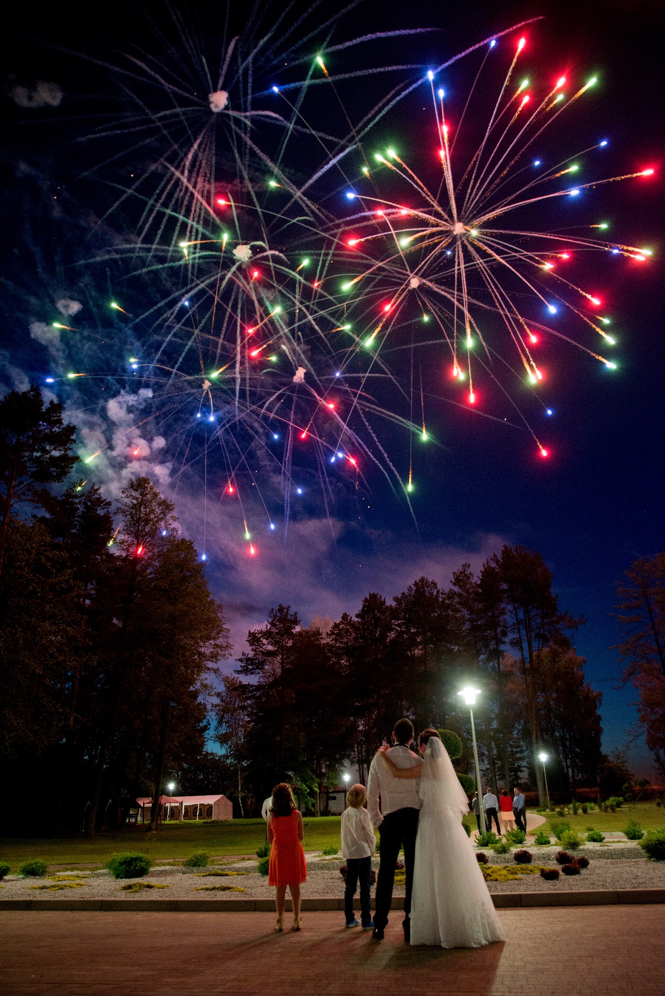 Para młoda i dzieci oglądają wielobarwne fajerwerki na tle nocnego nieba podczas uroczystości ślubnej w plenerze.