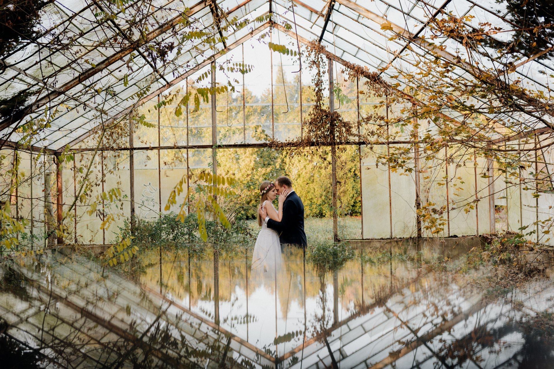 Para młoda obejmująca się w opuszczonej szklarni, z odbiciem konstrukcji w wodzie na podłodze i jesiennymi liśćmi na ścianach.