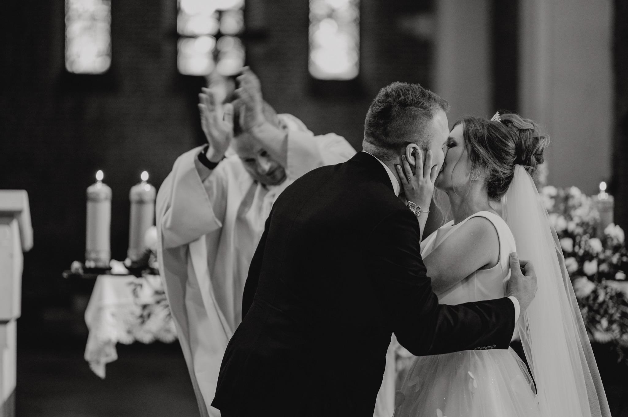 Czarno-białe zdjęcie nowożeńców całujących się w kościele w Zaleszanach, ksiądz błogosławi w tle, widoczne świece i kwiaty.