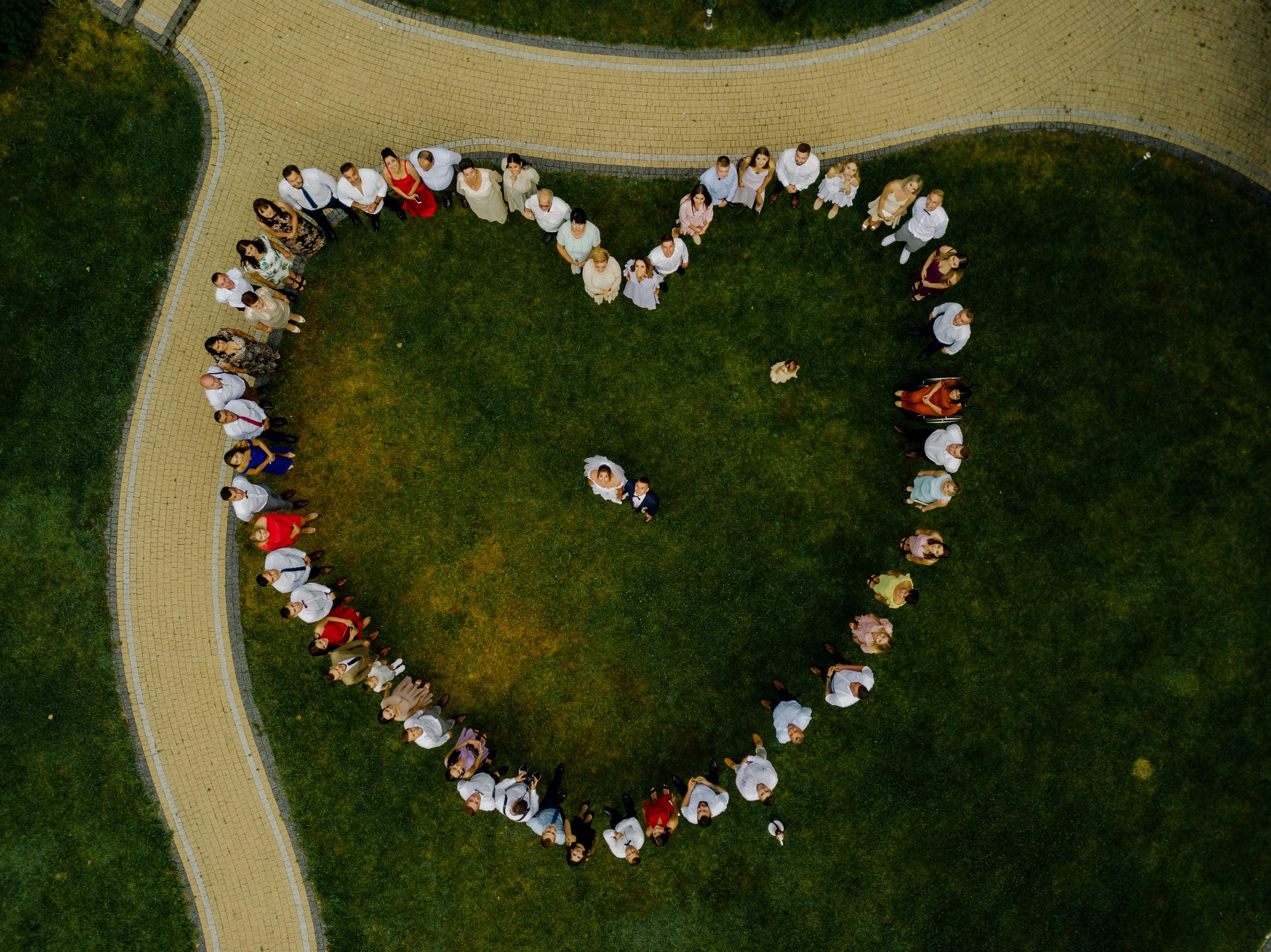 Widok z lotu ptaka na grupę ludzi ustawionych w kształt serca na trawniku, para młoda w centrum, obok ścieżka z kostki brukowej.