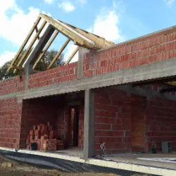 Surowy stan domu z czerwonej cegły z widoczną konstrukcją dachu i stertą cegieł wewnątrz, na tle błękitnego nieba z chmurami.